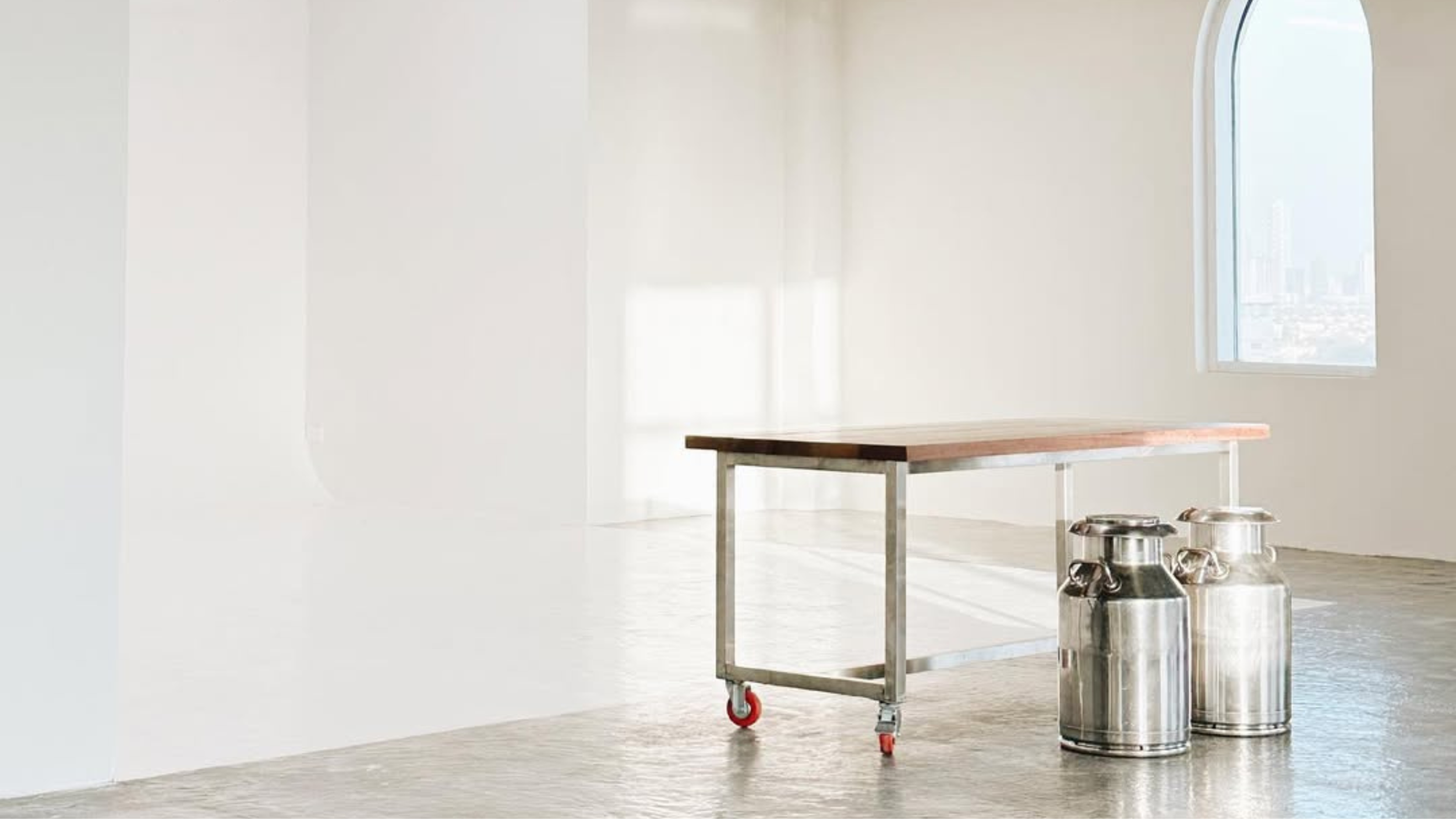 Empty room with white walls and large window, a wooden table on wheels, and stainless steel milk cans on the floor.
