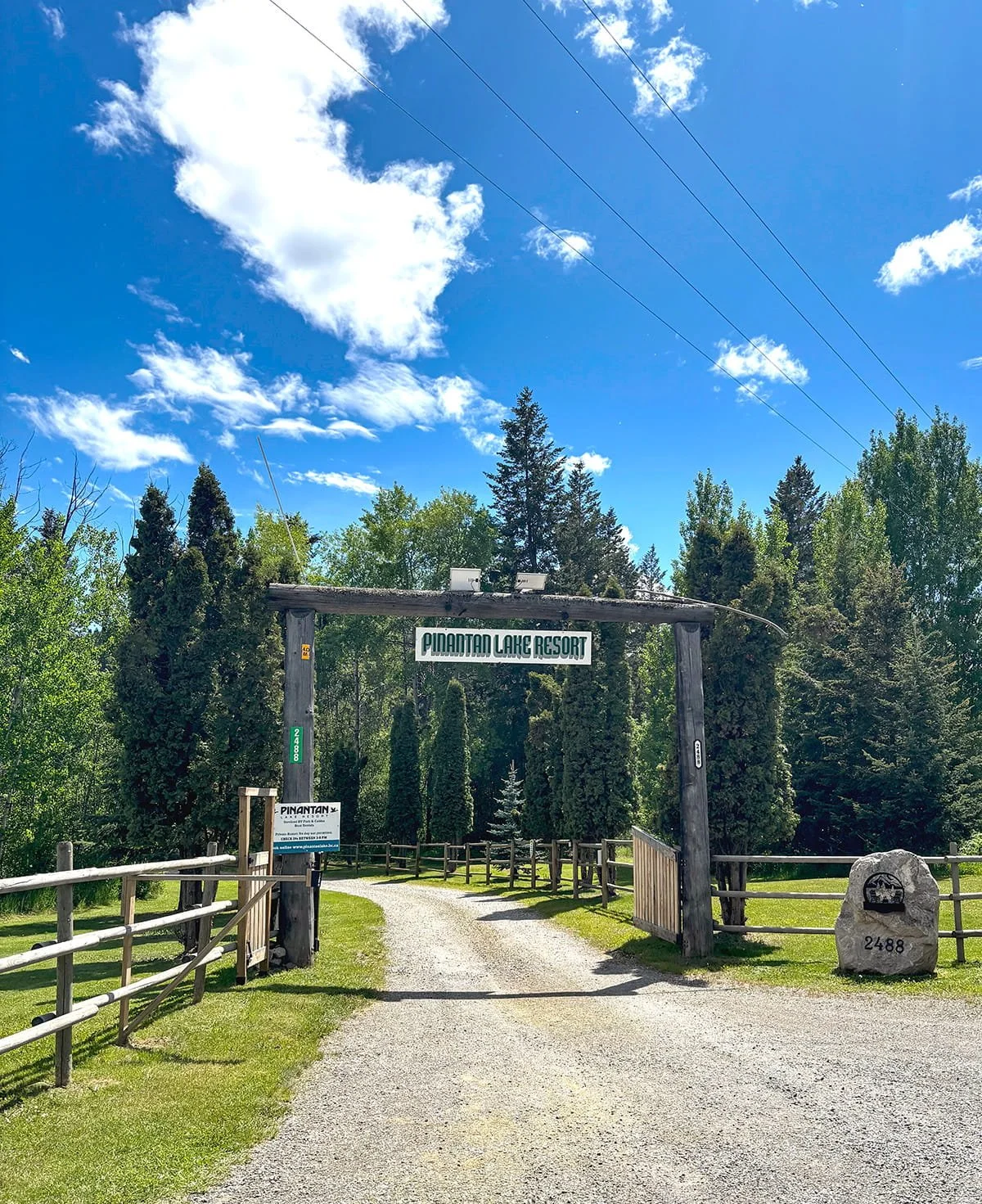 Entrance sign for Pinantan Lake Resort with a gravel driveway, surrounded by trees and a blue sky with clouds.