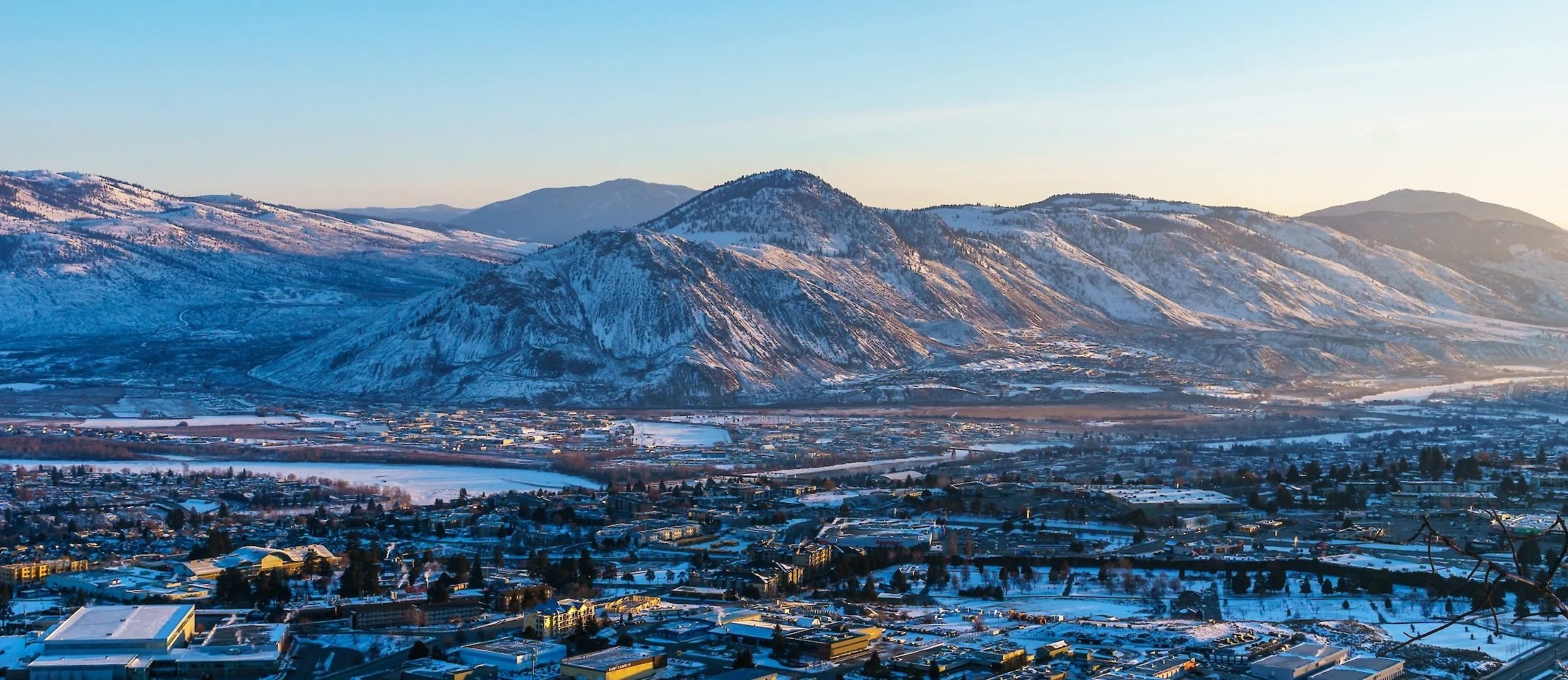 A snow-covered cityscape at the foot of snow-capped mountains during sunset.