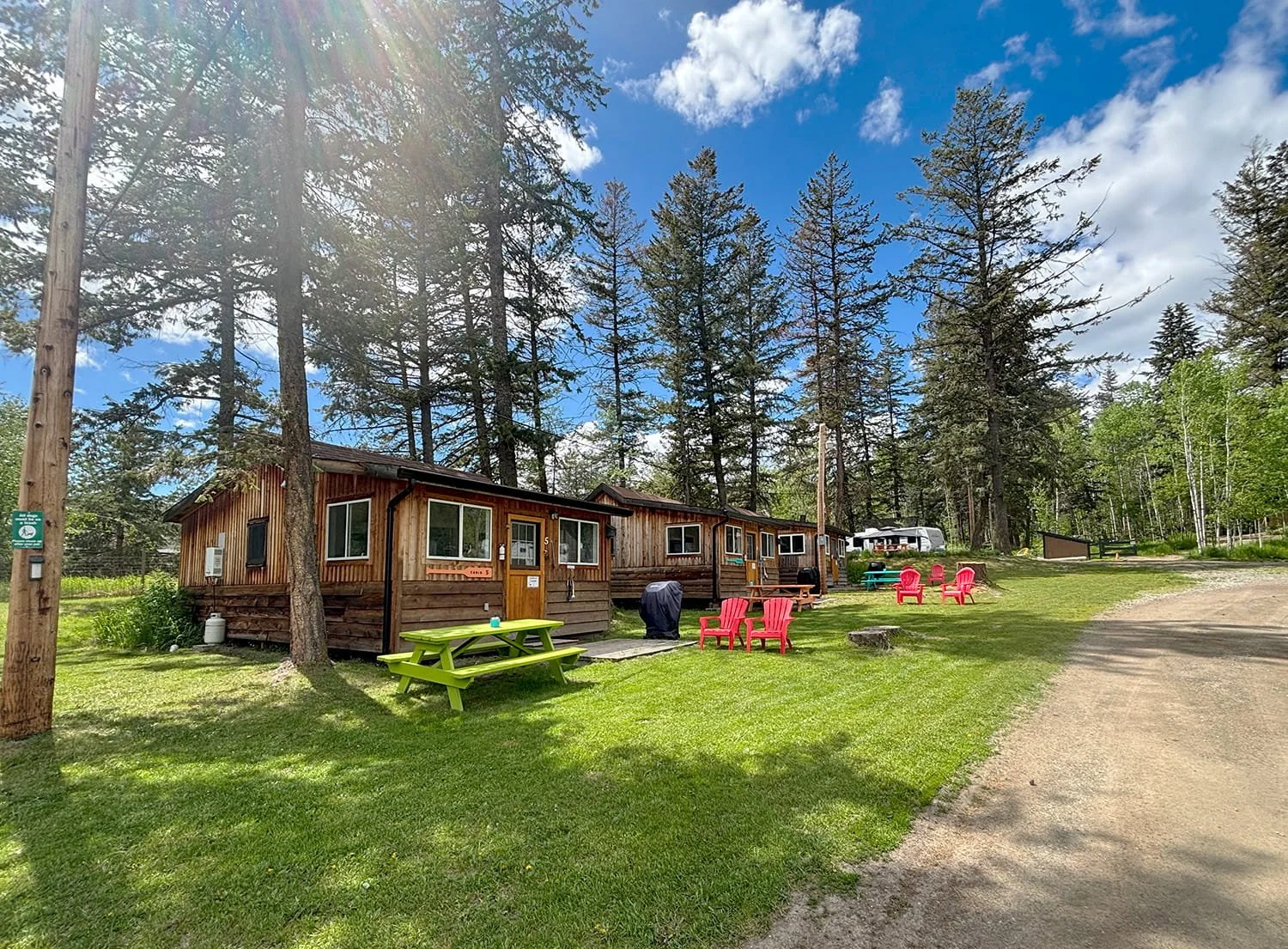Wooden cabins in a forested area with green grass, colorful Adirondack chairs, a picnic table, and a gravel path, under a blue sky with scattered clouds.