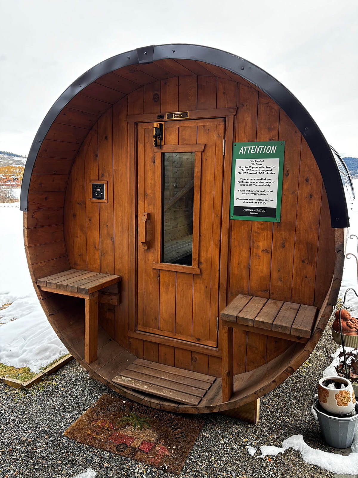 A round wooden sauna with a door and two small benches outside on a gravel surface, with snow on the ground and a cloudy sky in the background. There is a green sign on the sauna with safety instructions and a small black panel on the left side of the door.