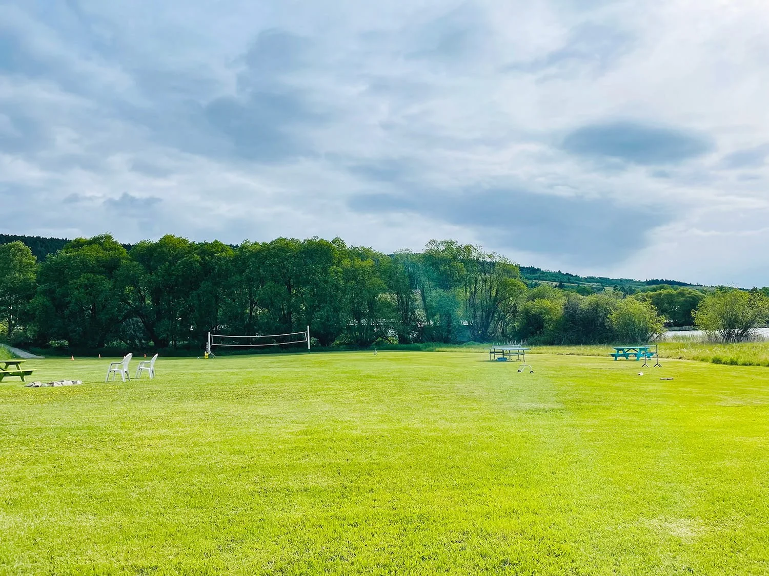 A grassy field with scattered picnic tables, chairs, and sports equipment under a partly cloudy sky, with trees and a lake in the background.