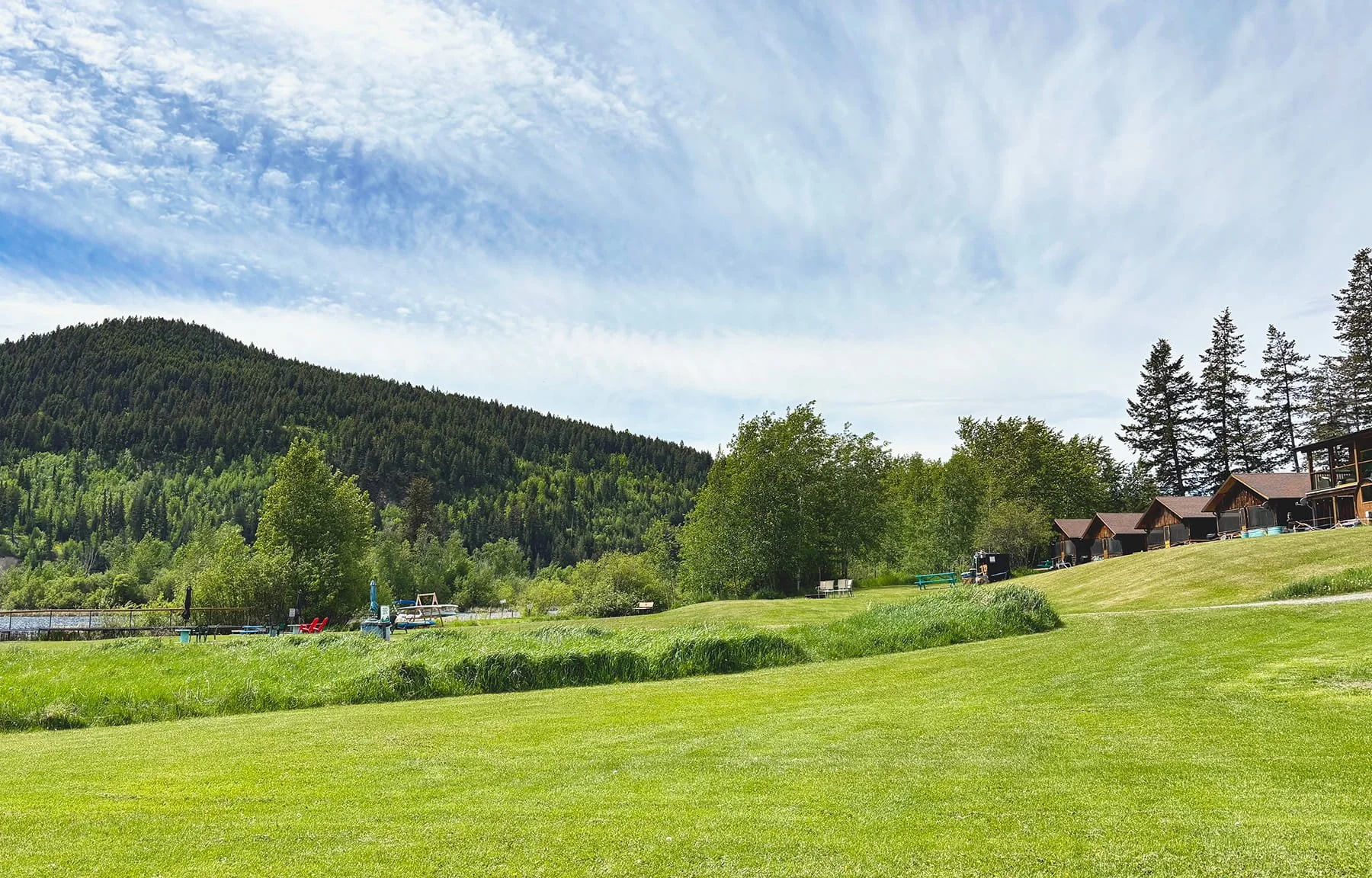 Scenic landscape of a lush green grassy area with trees, cottages, and a mountain in the background under a partly cloudy sky.