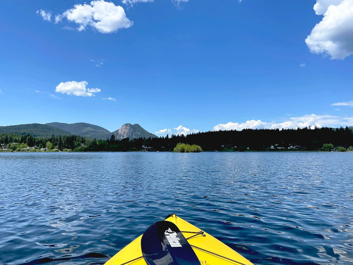 View from a yellow kayak on a calm lake, with a paddle resting on the kayak, surrounded by forested hills and mountains under a partly cloudy blue sky.
