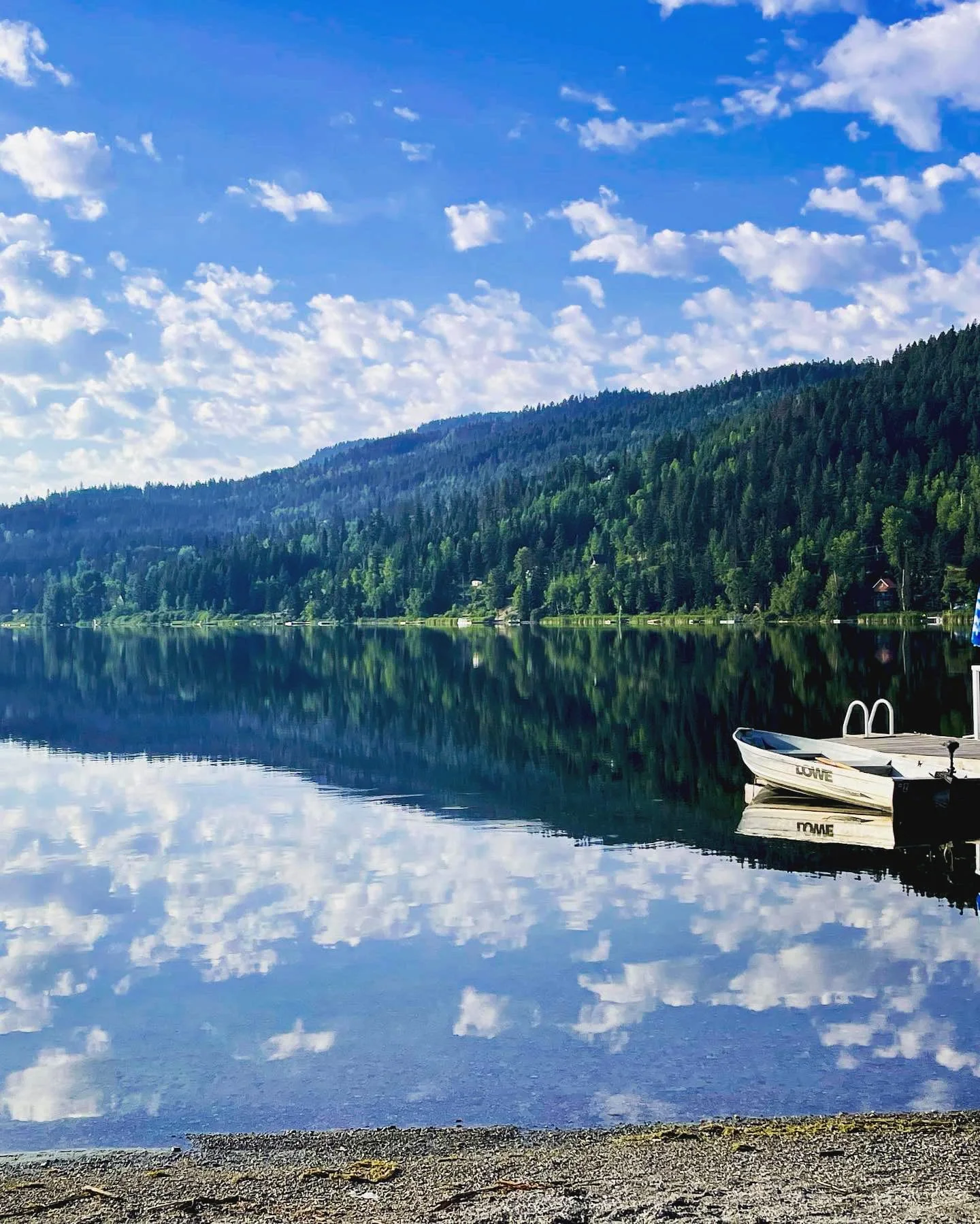 A peaceful lakeside scene with a mountain and tree-covered hill reflected in the calm water, and a small white boat docked at the shore under a partly cloudy sky.