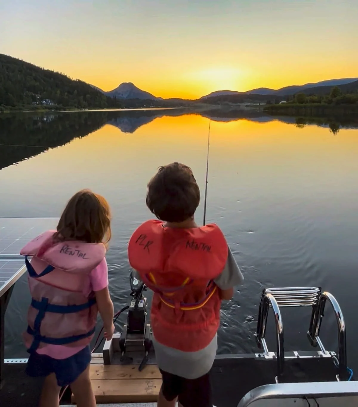 Two children, one girl and one boy, standing on the edge of a boat, fishing at sunset on a calm lake with mountains in the background.
