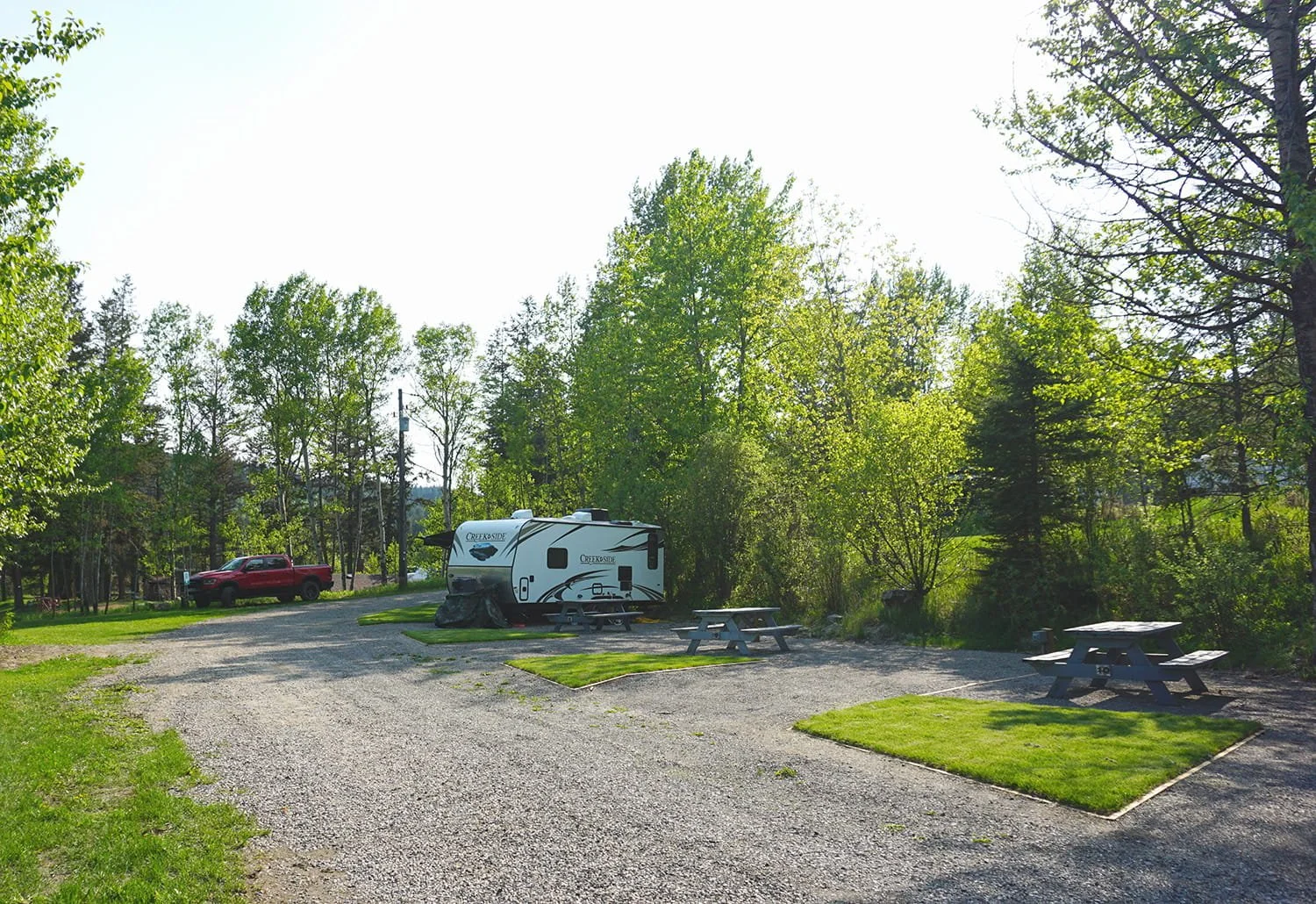 A campground with a gravel parking area, a recreational vehicle, a red pickup truck, wooden picnic tables, and surrounded by green trees.