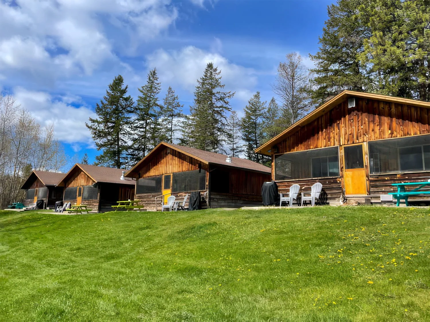 Three wooden cabins with screened porches on a grassy hill, surrounded by trees under a partly cloudy sky.