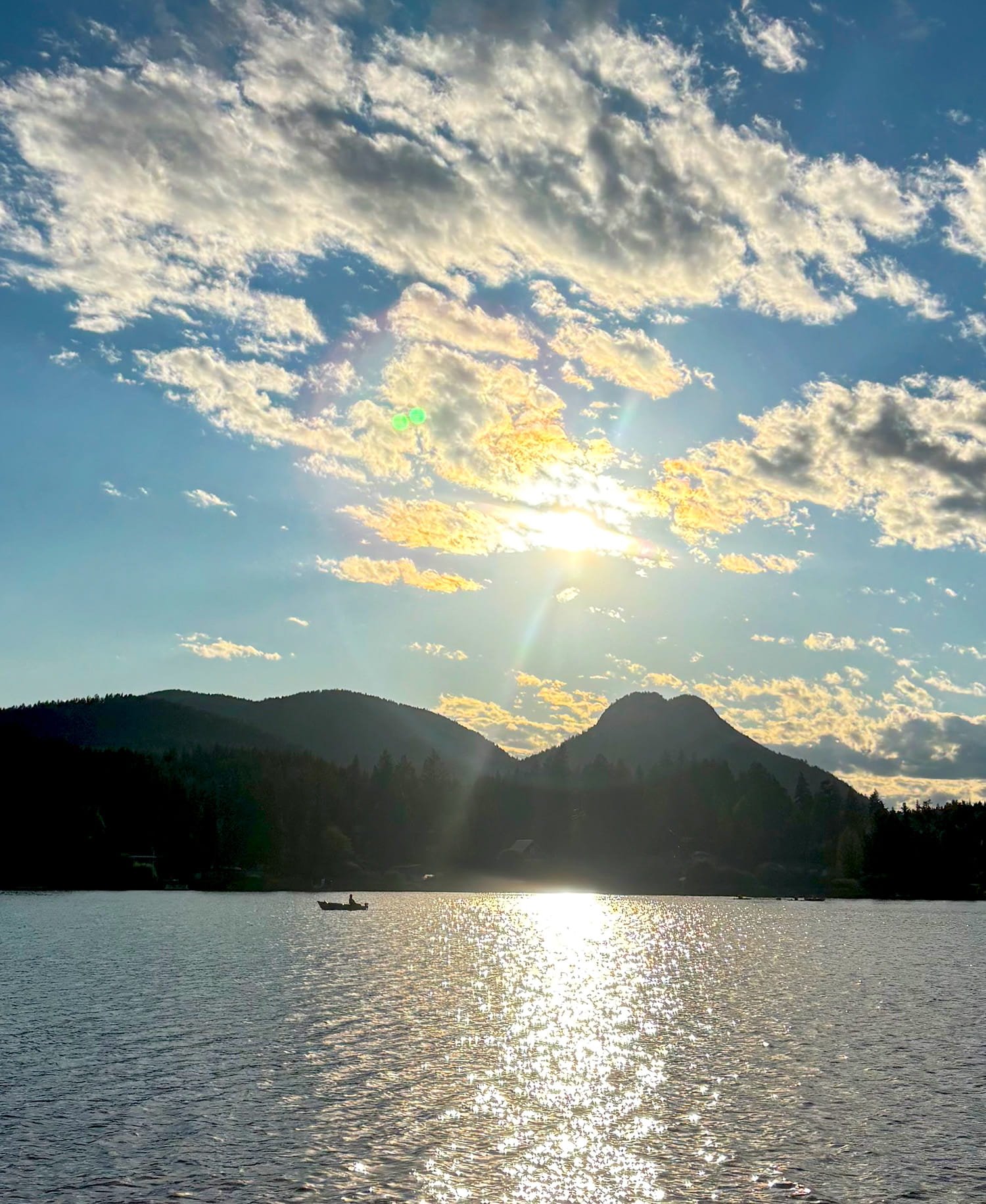 Sunset over a lake with mountains in the background, a boat on the water, and partly cloudy sky with sunlight shining through.