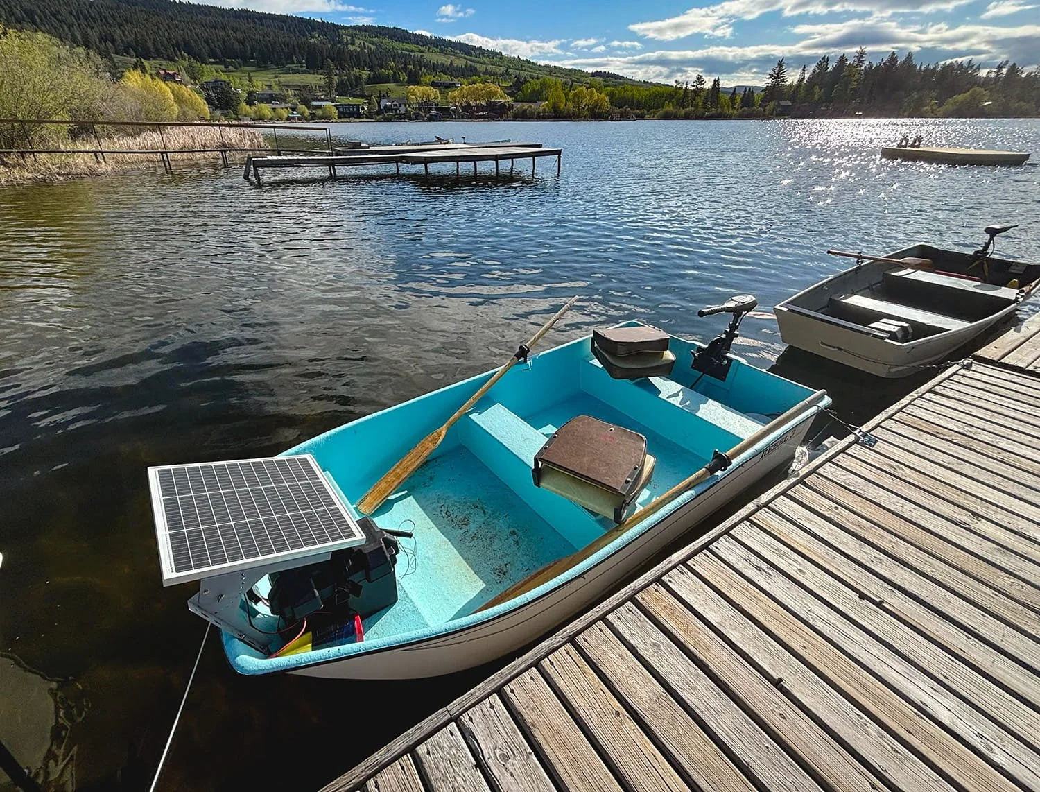 A small rowboat with a solar panel, paddle, and other equipment docked at a wooden pier on a lake, with trees, hills, and houses in the background on a bright sunny day.