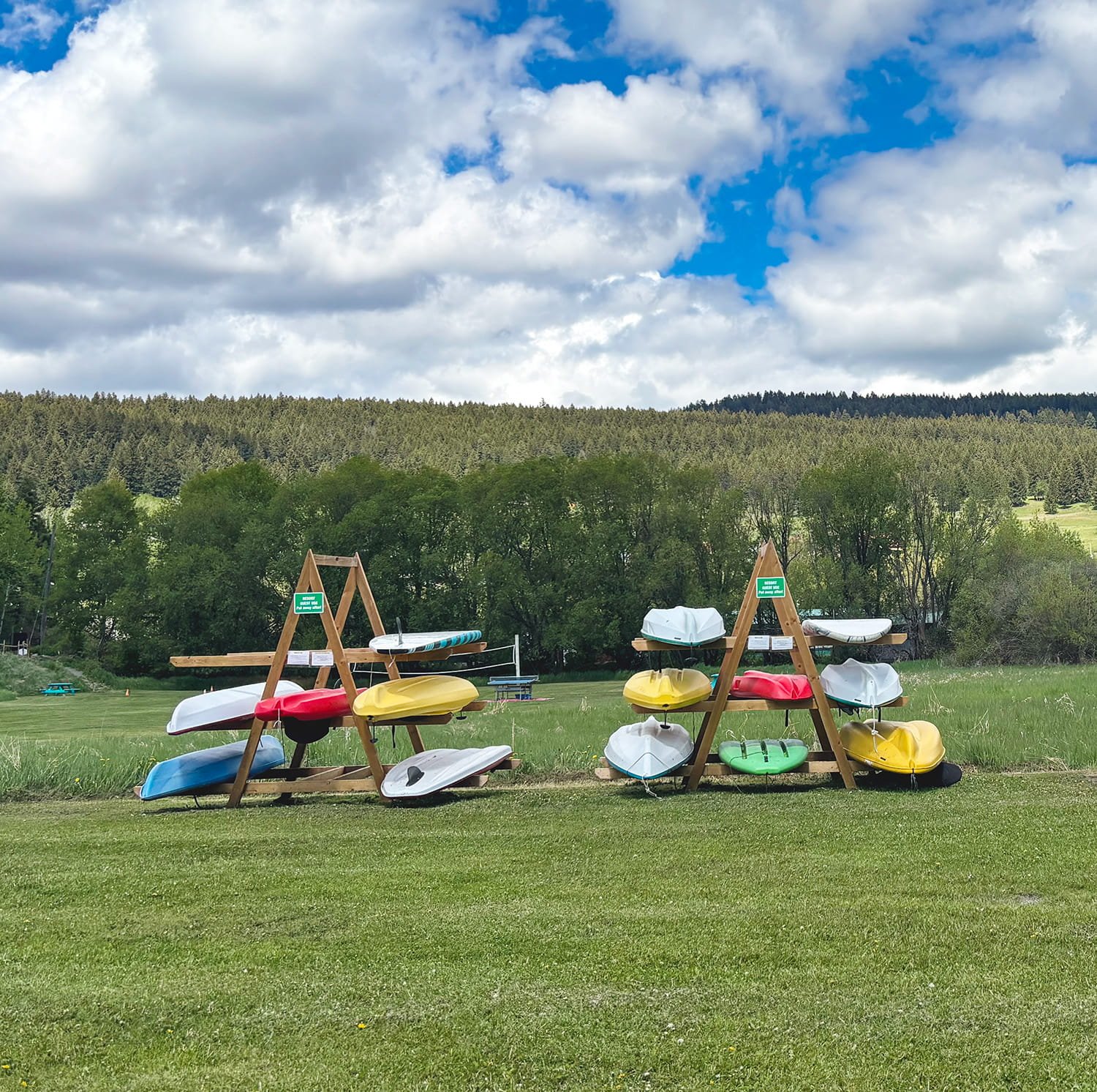 Multiple colorful kayaks stored on wooden racks in a grassy outdoor area, with trees and hills in the background under a partly cloudy sky.