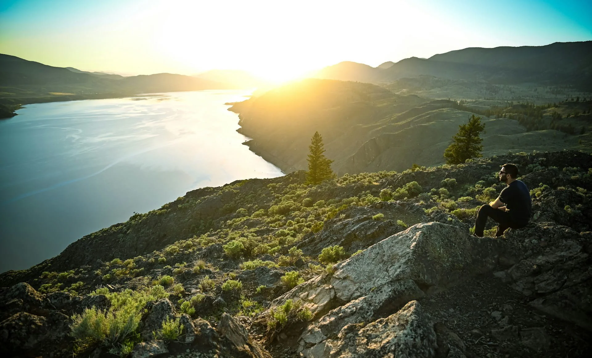 A person sitting on rocks looking over a large lake with mountains in the background during sunset.