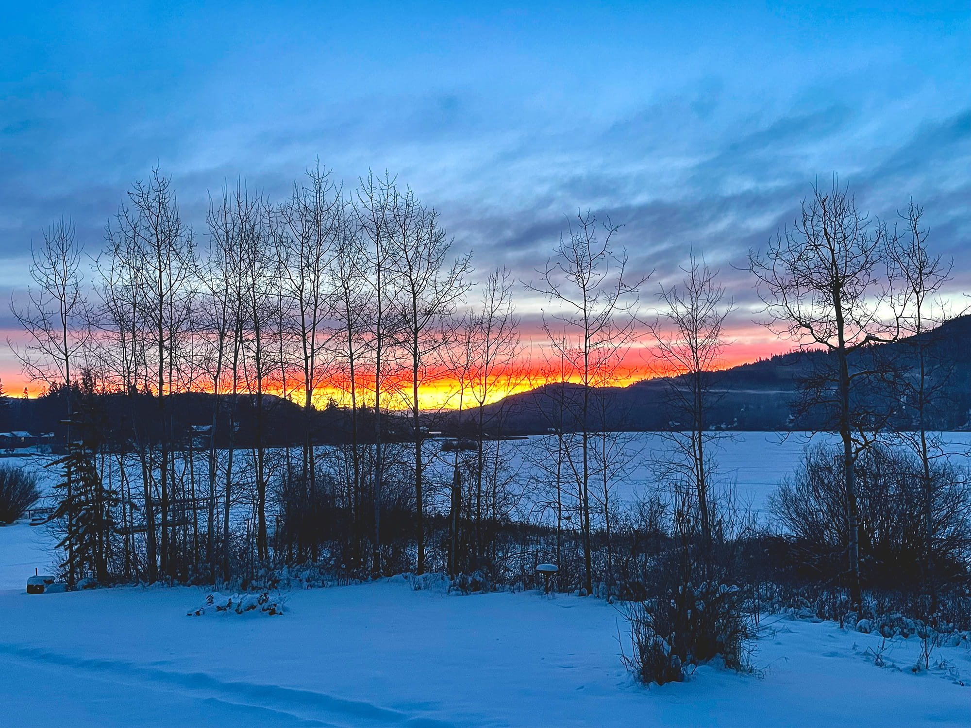 Snow-covered landscape with leafless trees in the foreground, a frozen lake, and distant mountains under a colorful sunset sky with shades of orange, pink, and blue.
