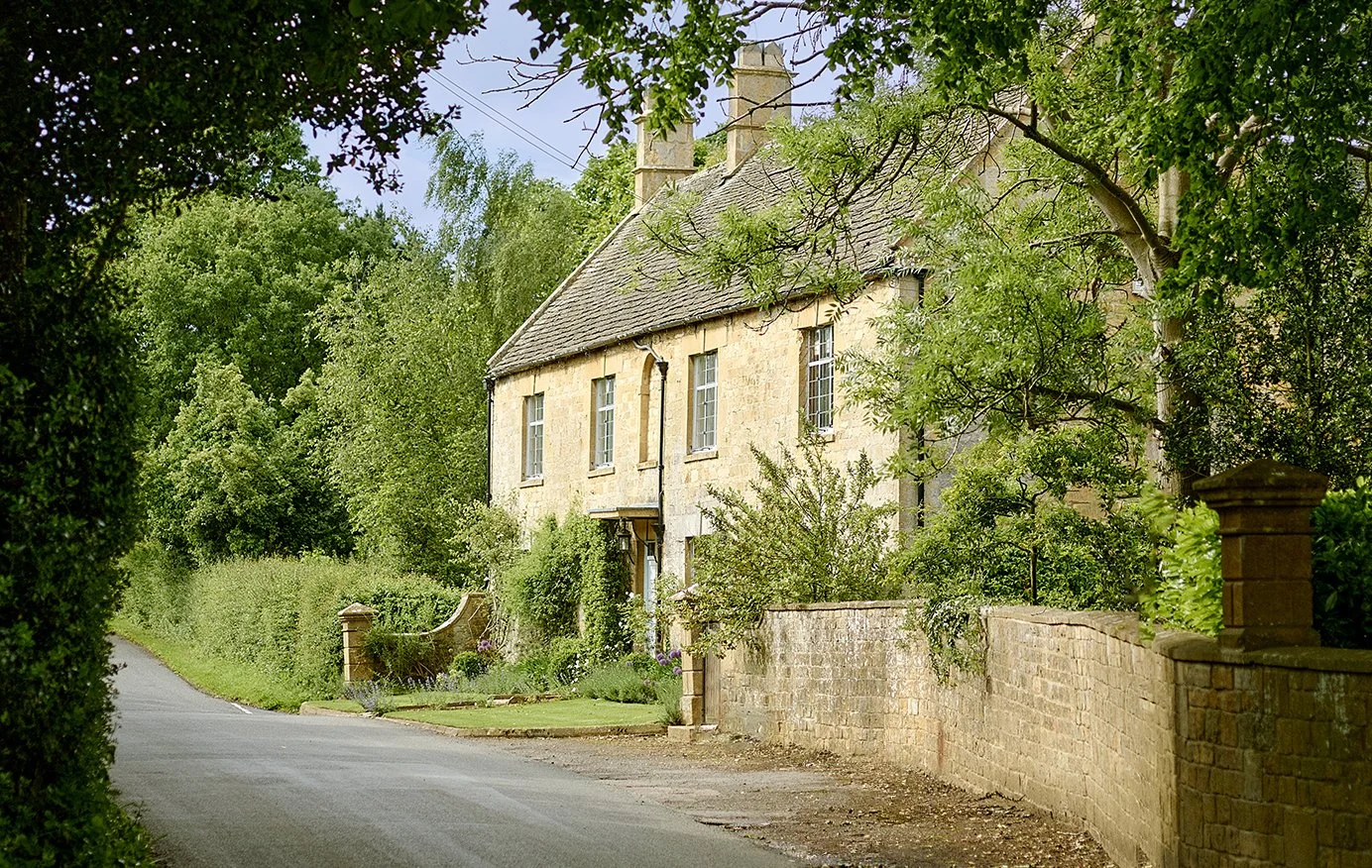 A scenic country house with a stone exterior, tall windows, and a sloped roof, surrounded by lush green trees and bushes, situated along a winding road.