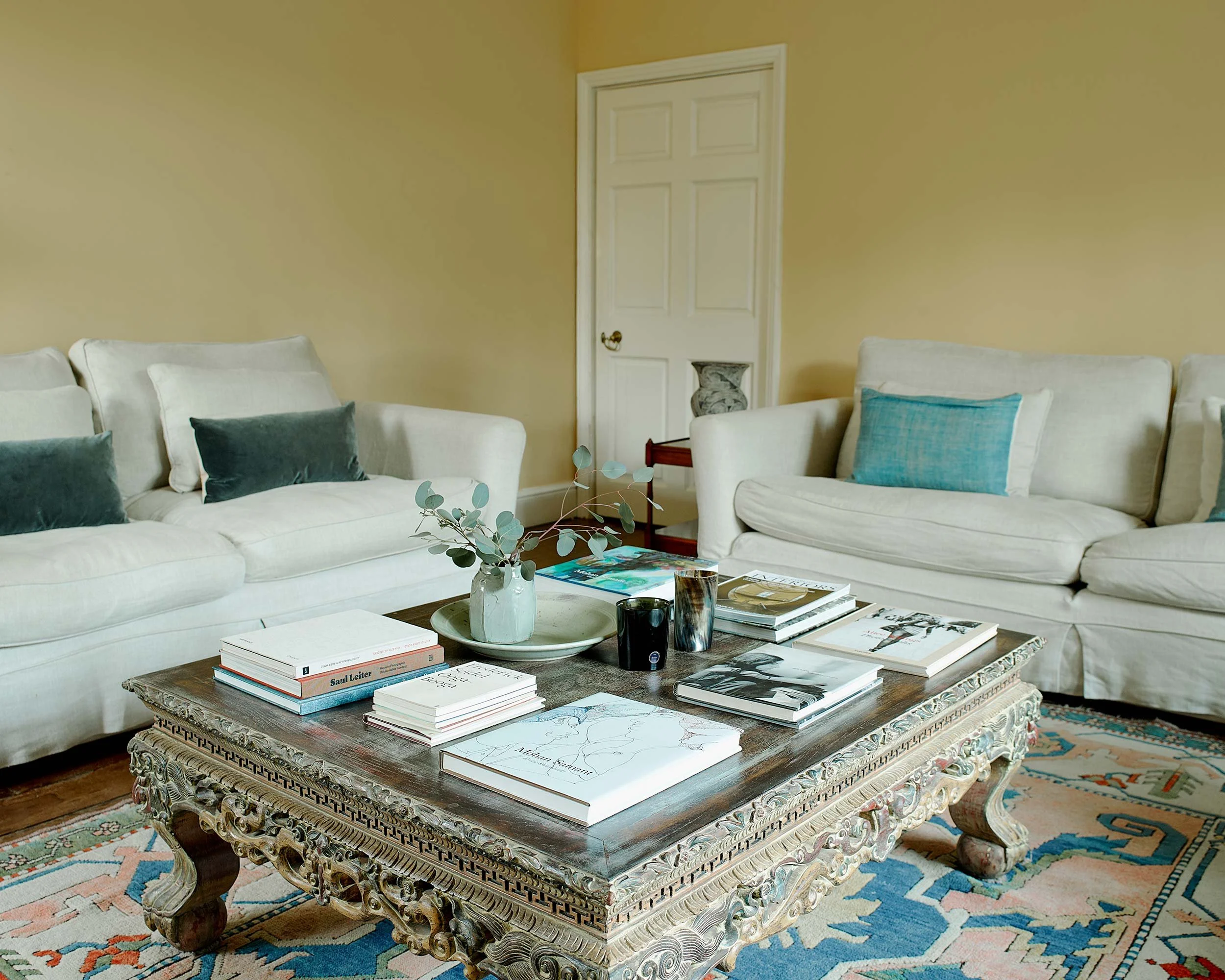 Living room with cream sofas, colorful pillows, a wooden coffee table covered with books and magazines, a potted plant, and a patterned area rug, with a yellow wall and white door in the background.