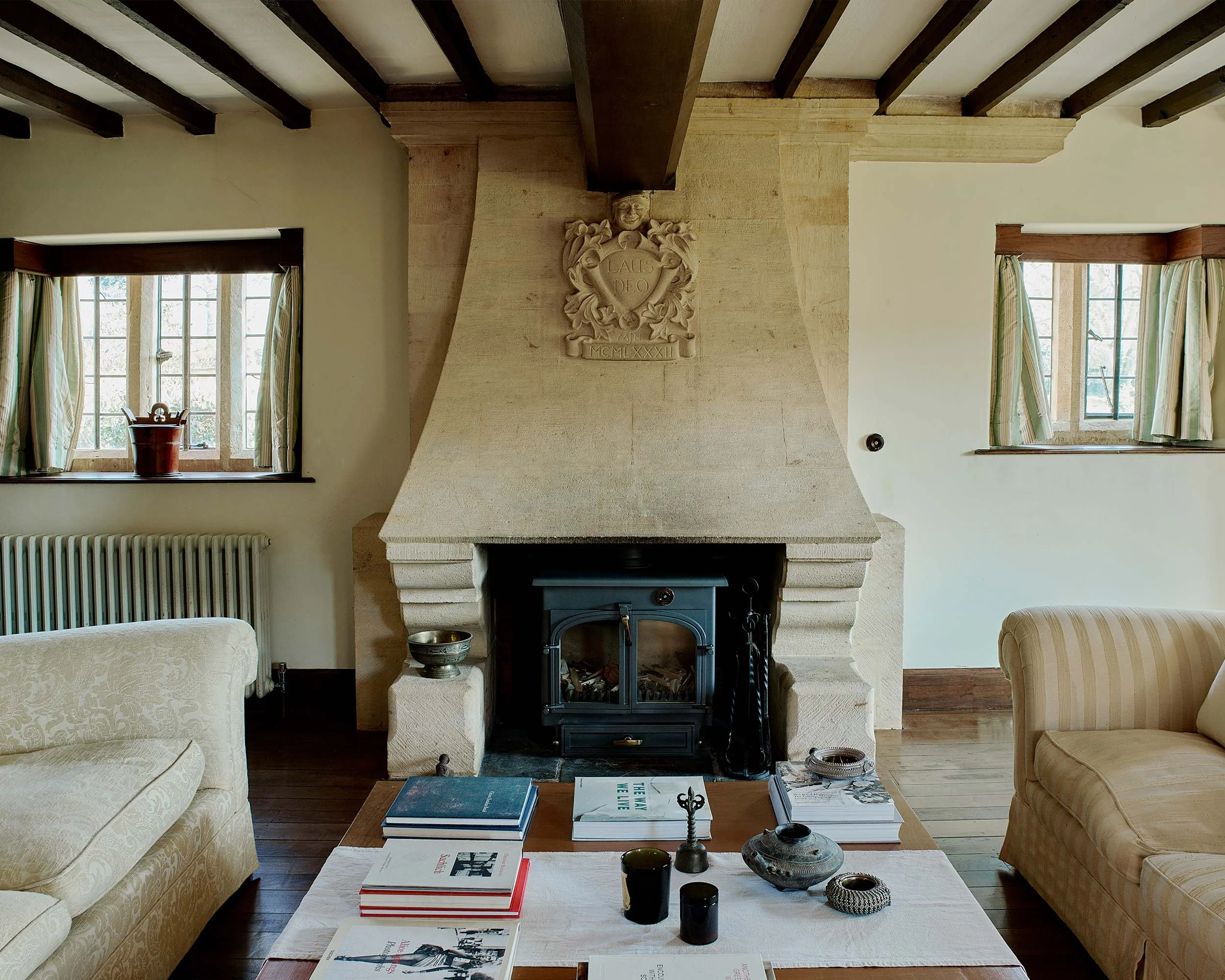 Living room with wood-beamed ceiling, beige sofas, a fireplace with decorative carving, and a wooden coffee table with books and decorative items, large windows with green curtains.