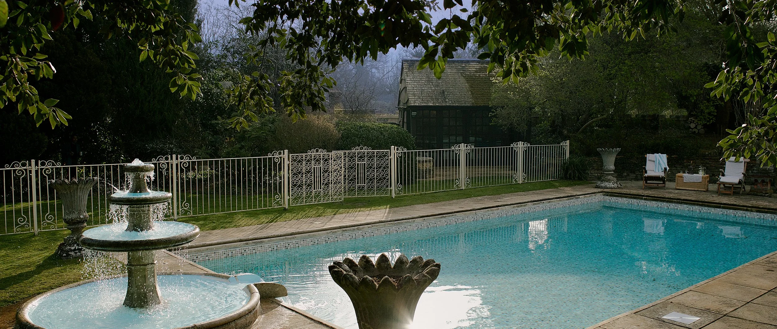 A backyard with a swimming pool, a two-tier fountain with water flowing, white metal fence, chairs with towels, and lush greenery.