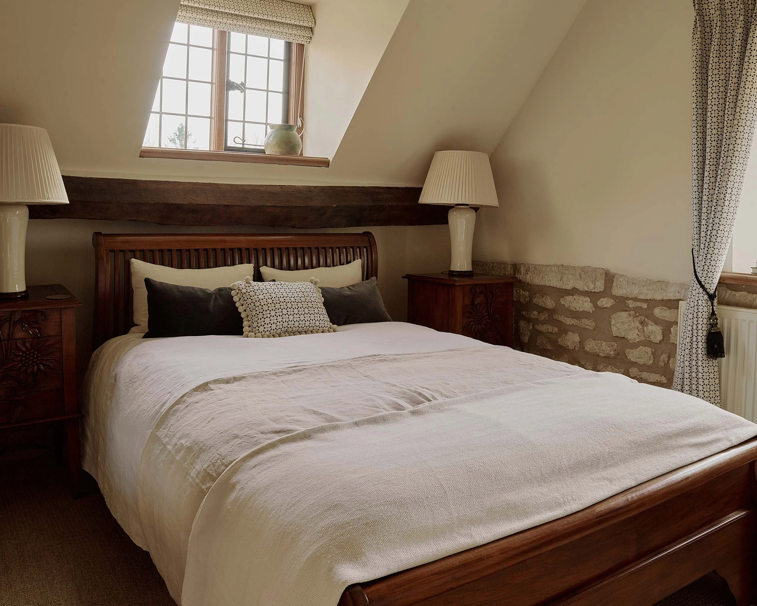 A cozy bedroom with a bed featuring a white textured bedspread, decorative pillows, two matching lamps on the nightstands, a small window with a polka dot curtain, and wooden beams and stone accents on the wall.