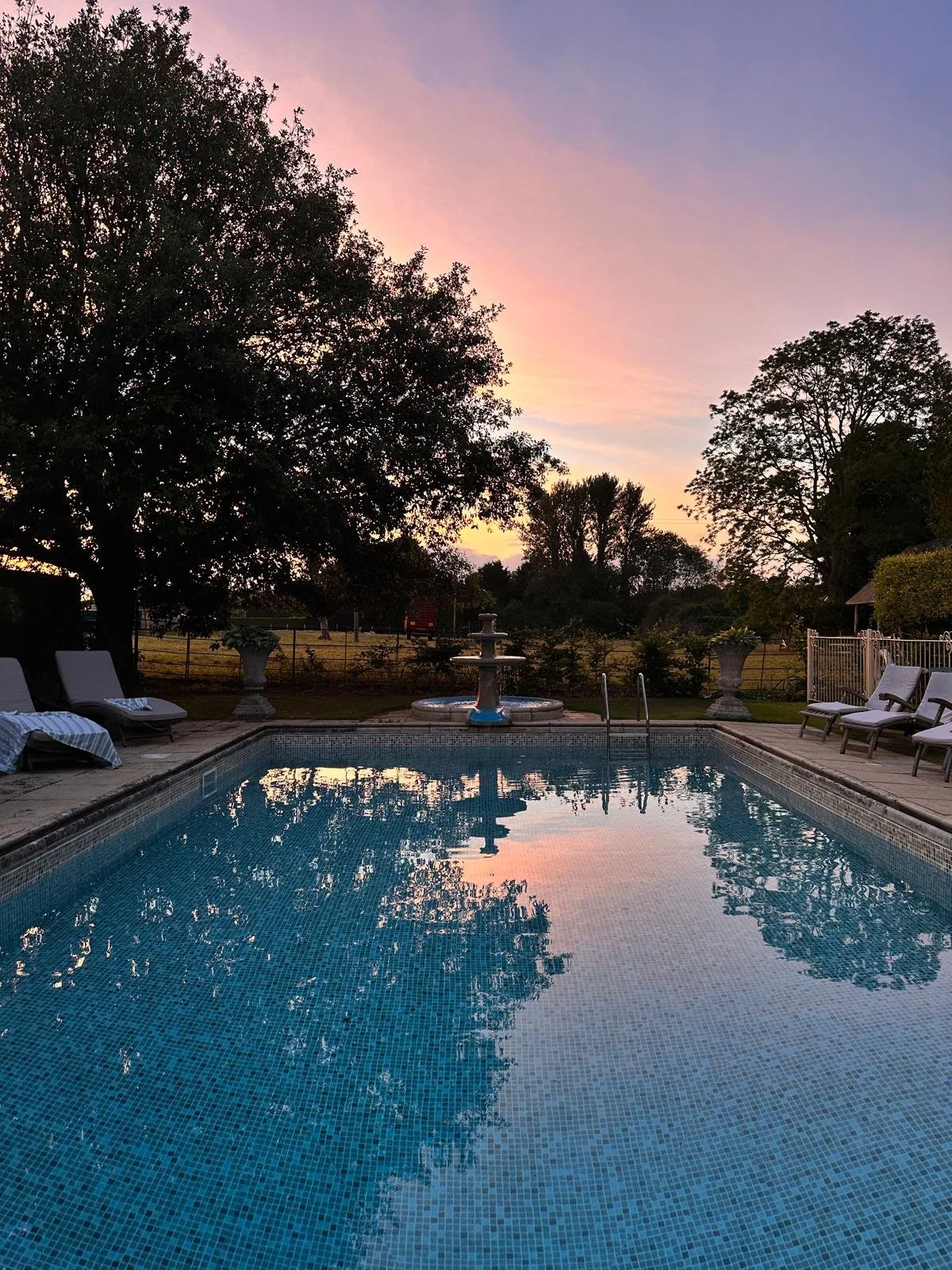 A backyard swimming pool at sunset with lounge chairs, trees, and a fountain, reflecting the colorful sky.