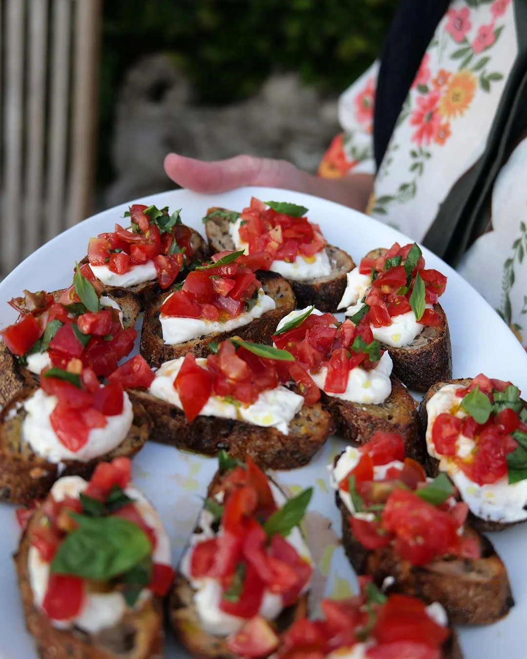 A person holding a white plate with toasted bread slices topped with mozzarella cheese, chopped tomatoes, and fresh basil.