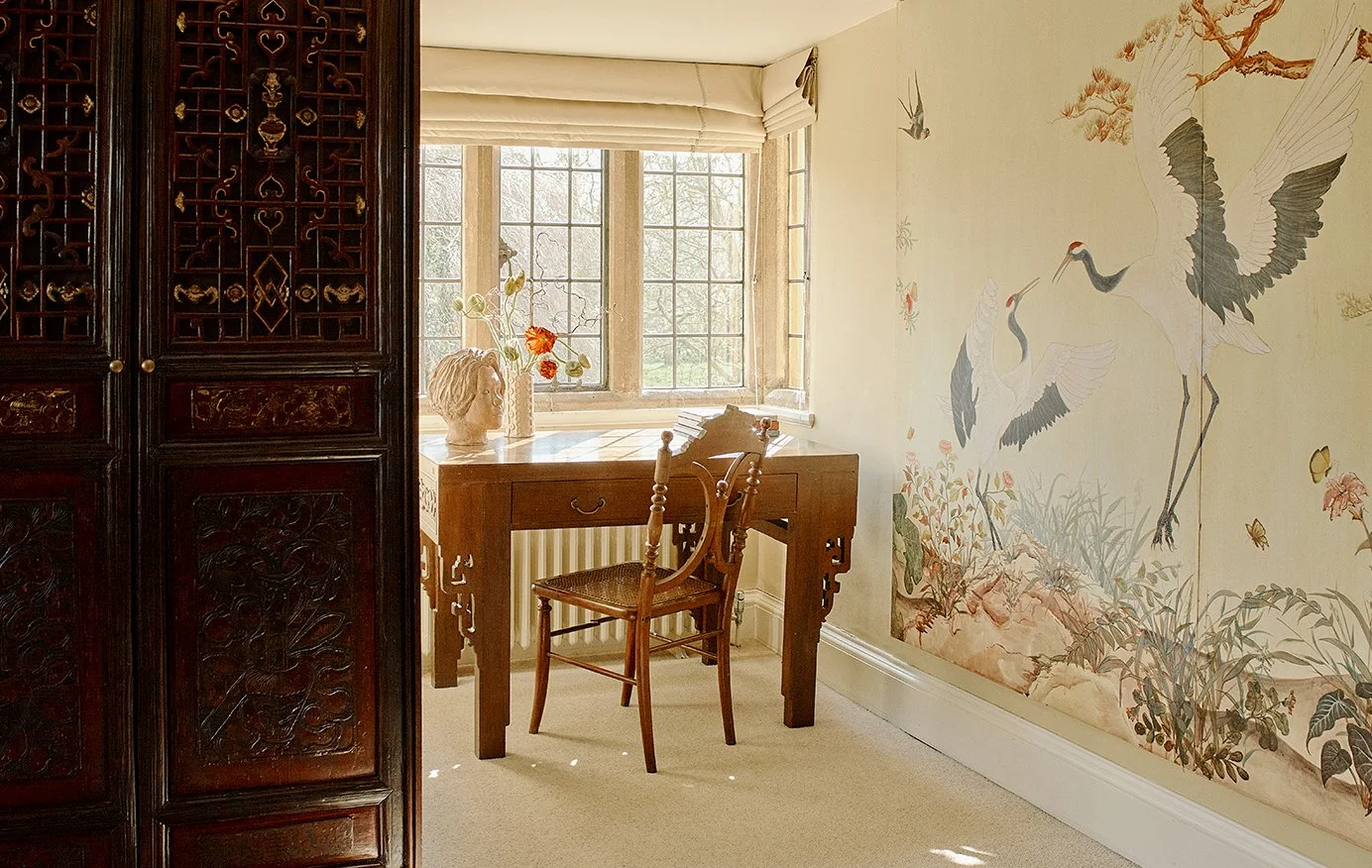 A cozy room with a wooden desk and chair near a large window, decorated with a bust sculpture and a vase of flowers, and featuring a mural of cranes and butterflies on the wall.