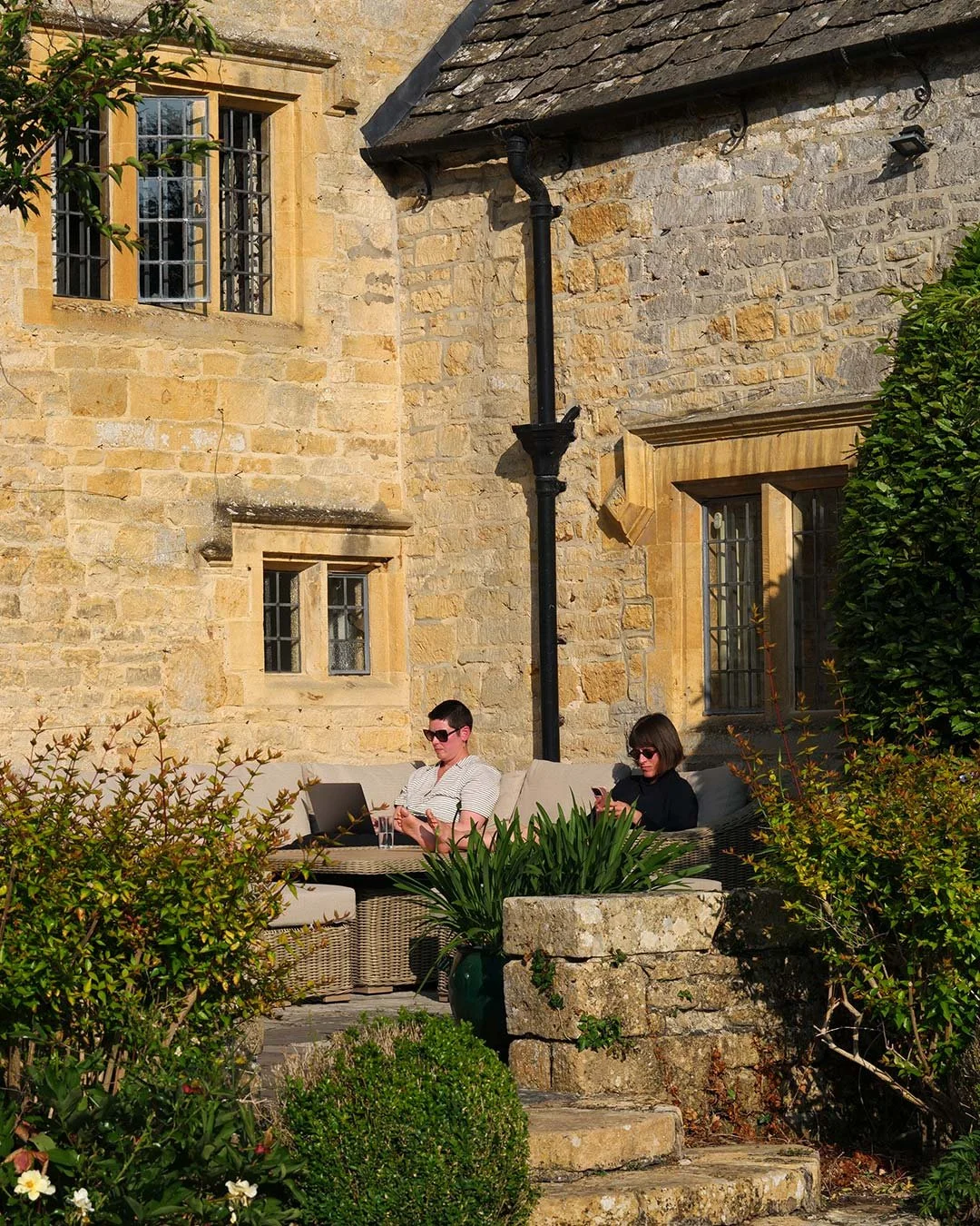 Two women sitting on outdoor cushions in a garden area with plants and stone steps, next to a building with stone walls and windows, one using a laptop and the other using a phone.