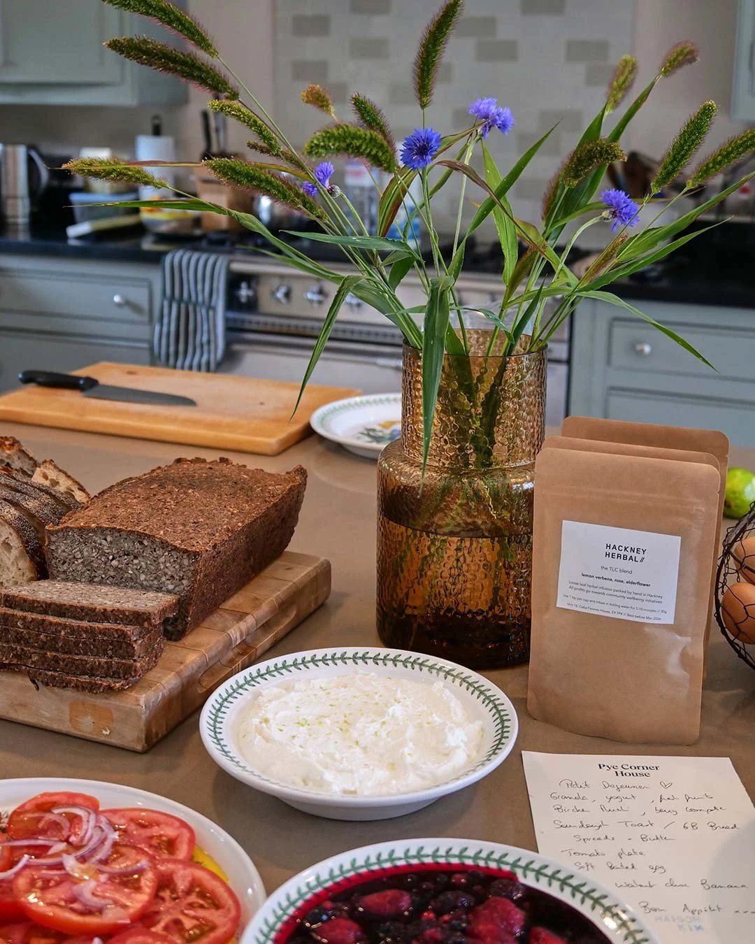 A kitchen counter with a loaf of bread, sliced and placed on a wooden cutting board, a bowl of cream cheese, a plate of sliced tomatoes with onions, a bowl of mixed berries, a bag of herbal tea labeled 'Hackney Herbal,' a vase with purple and green f