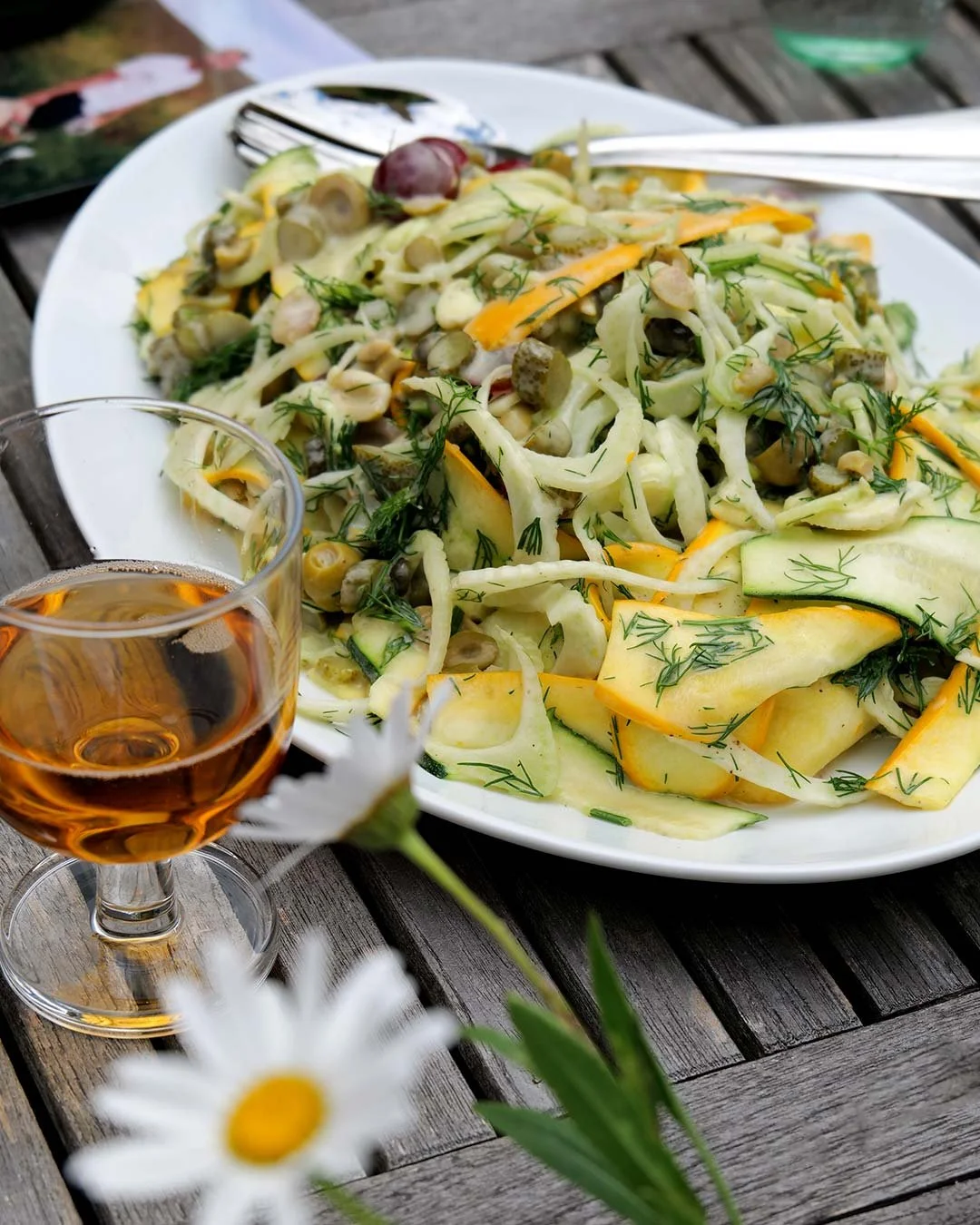 A salad with sliced vegetables, capers, and herbs on a white plate, accompanied by a glass of iced tea on a wooden table.