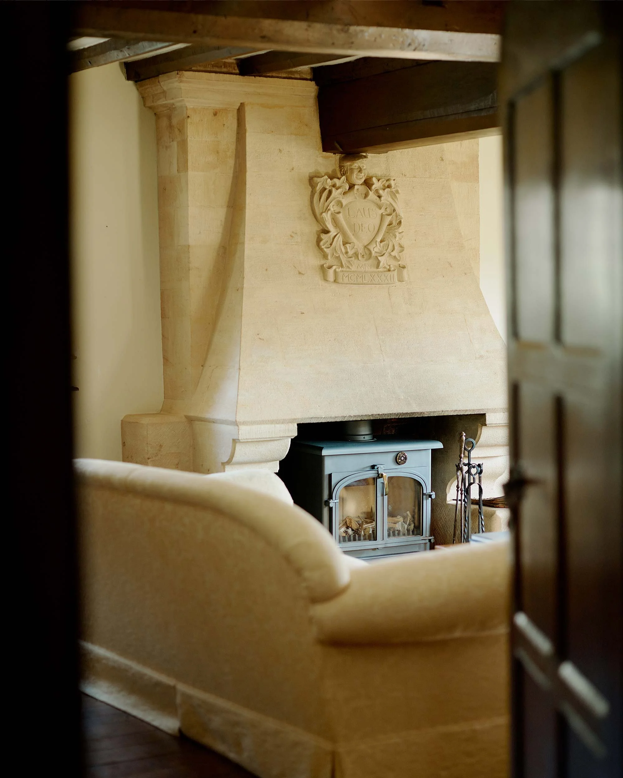 Looking through a small window or gap, viewing a living room with a beige couch, a fireplace with a metal glass door, and a wooden mantelpiece. On the wood wall above the fireplace, there is a decorative plaque with a shield, a smiling face, and insc