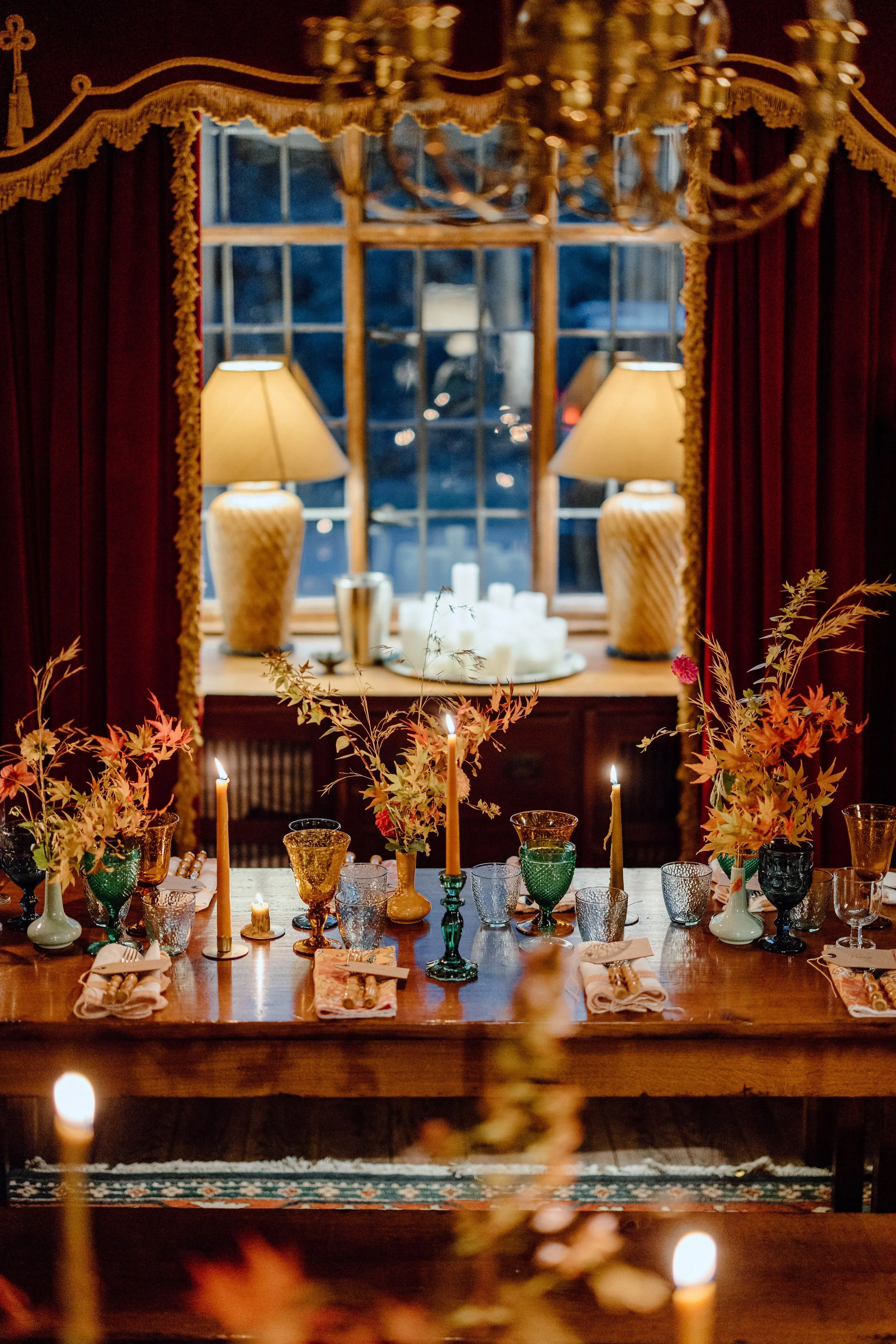 A dining table set with various colored glassware, candles, and floral arrangements, viewed from across the room with a window and lamps in the background.
