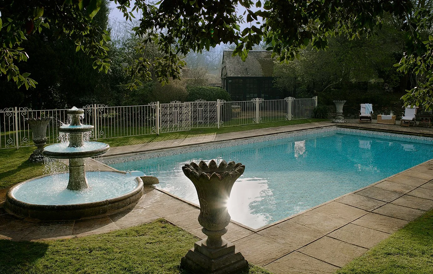 A backyard swimming pool with a fountain on the left and lounge chairs on the right, surrounded by a white metal fence and lush green trees.