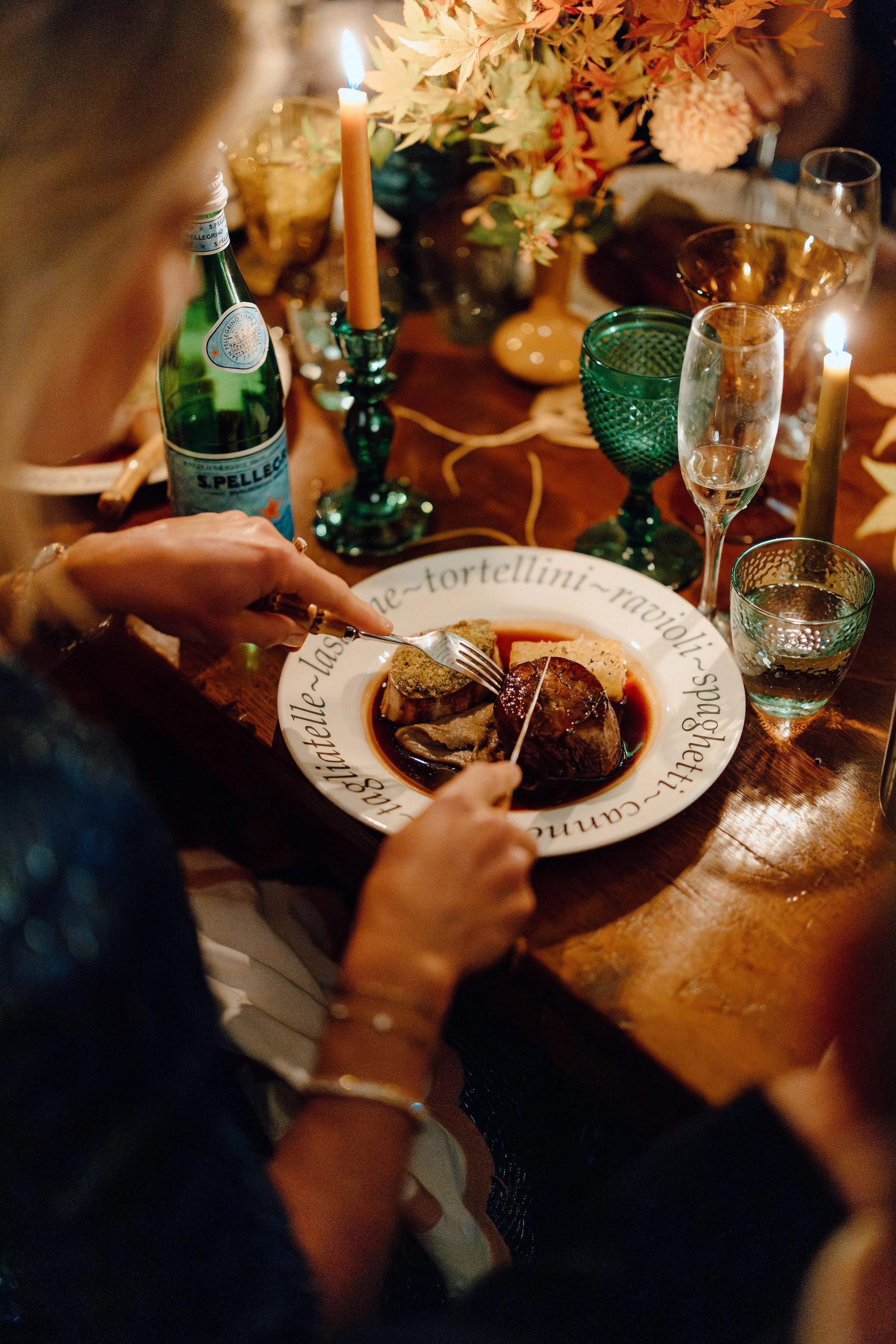 A dinner table setting with a plate of food, wine glasses, candles, and floral centerpieces, viewed from above and partially obscured by a person's arm and hand.