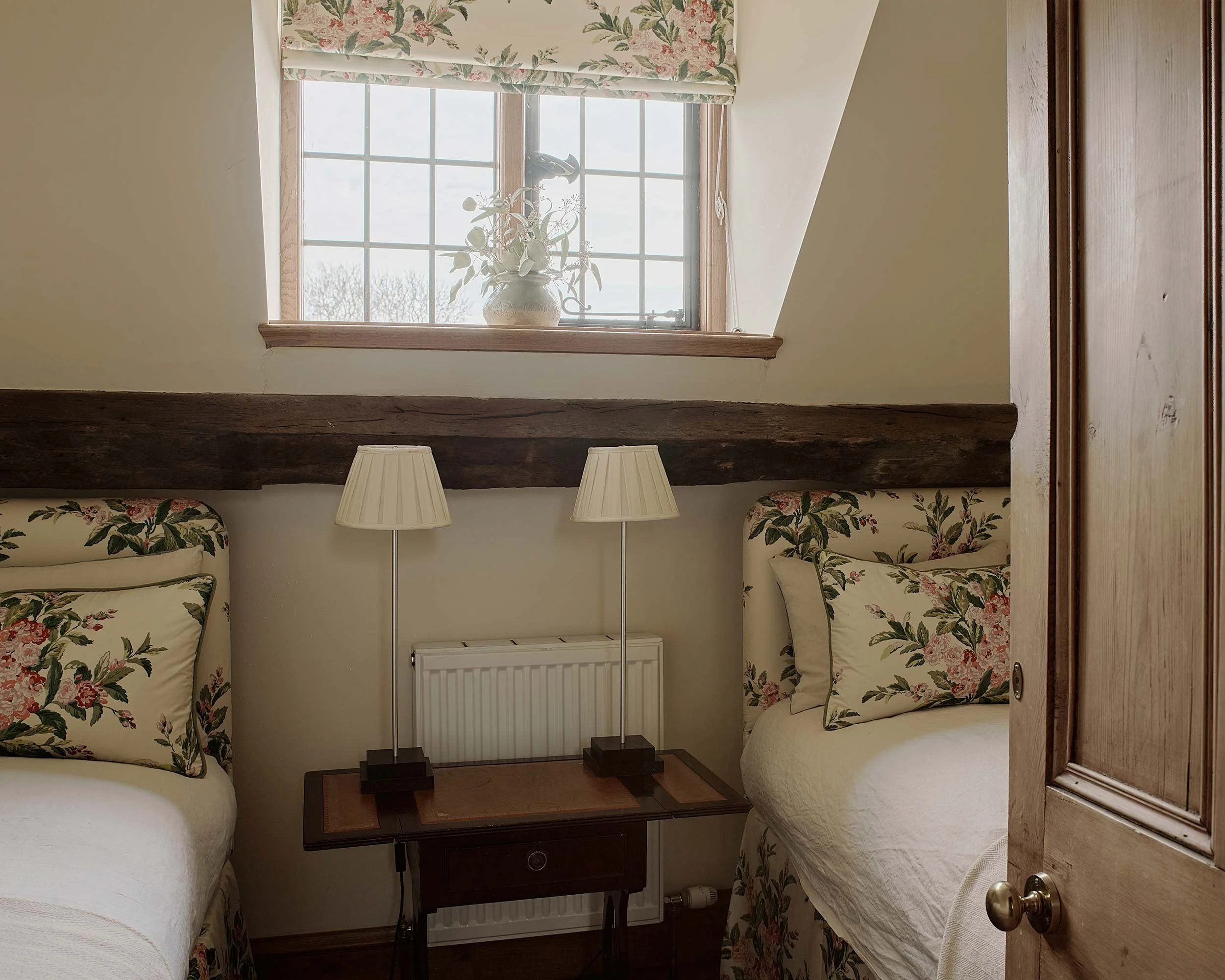 A cozy bedroom with two beds with floral-patterned bedding, a window with a floral valance, a central wooden table, and two lamps on either side of the table. The window has a plant in a decorative vase.