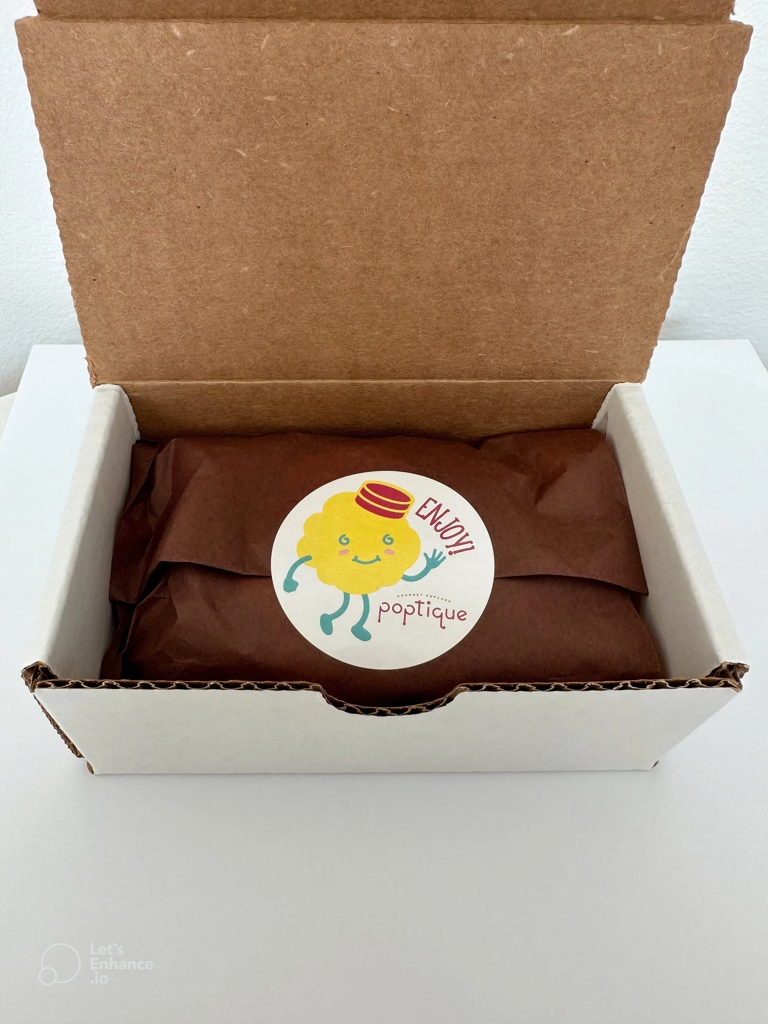Open white box with brown paper-wrapped item inside, featuring a circular sticker with a cartoon character and the words 'Enjoy' and 'poptique'.