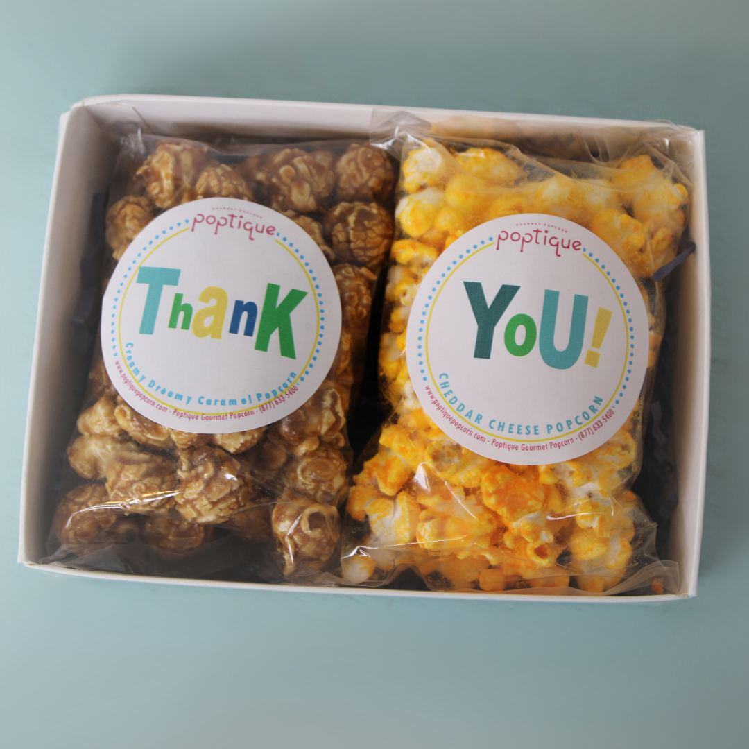 A white box containing two packs of gourmet popcorn, one caramel and the other cheddar cheese, each labeled with a colorful sticker saying "Thank" and "You!" respectively.