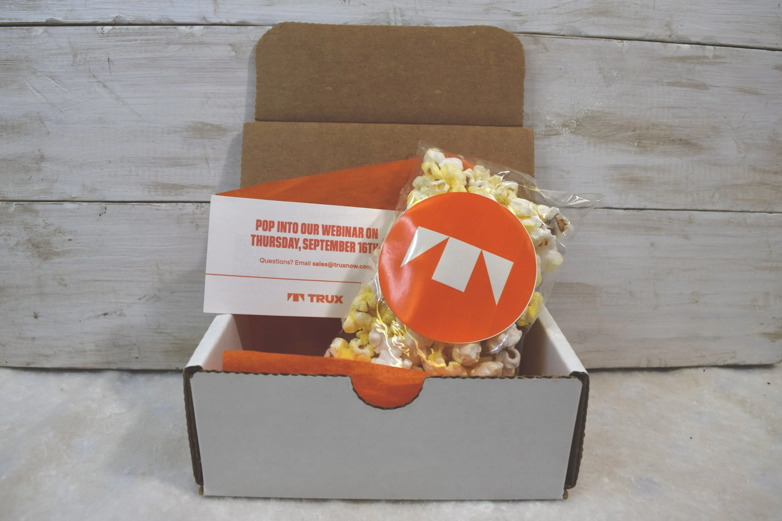 A white cardboard box containing a small bag of popcorn with a red and white logo, and a flyer advertising a webinar scheduled for September 16th.