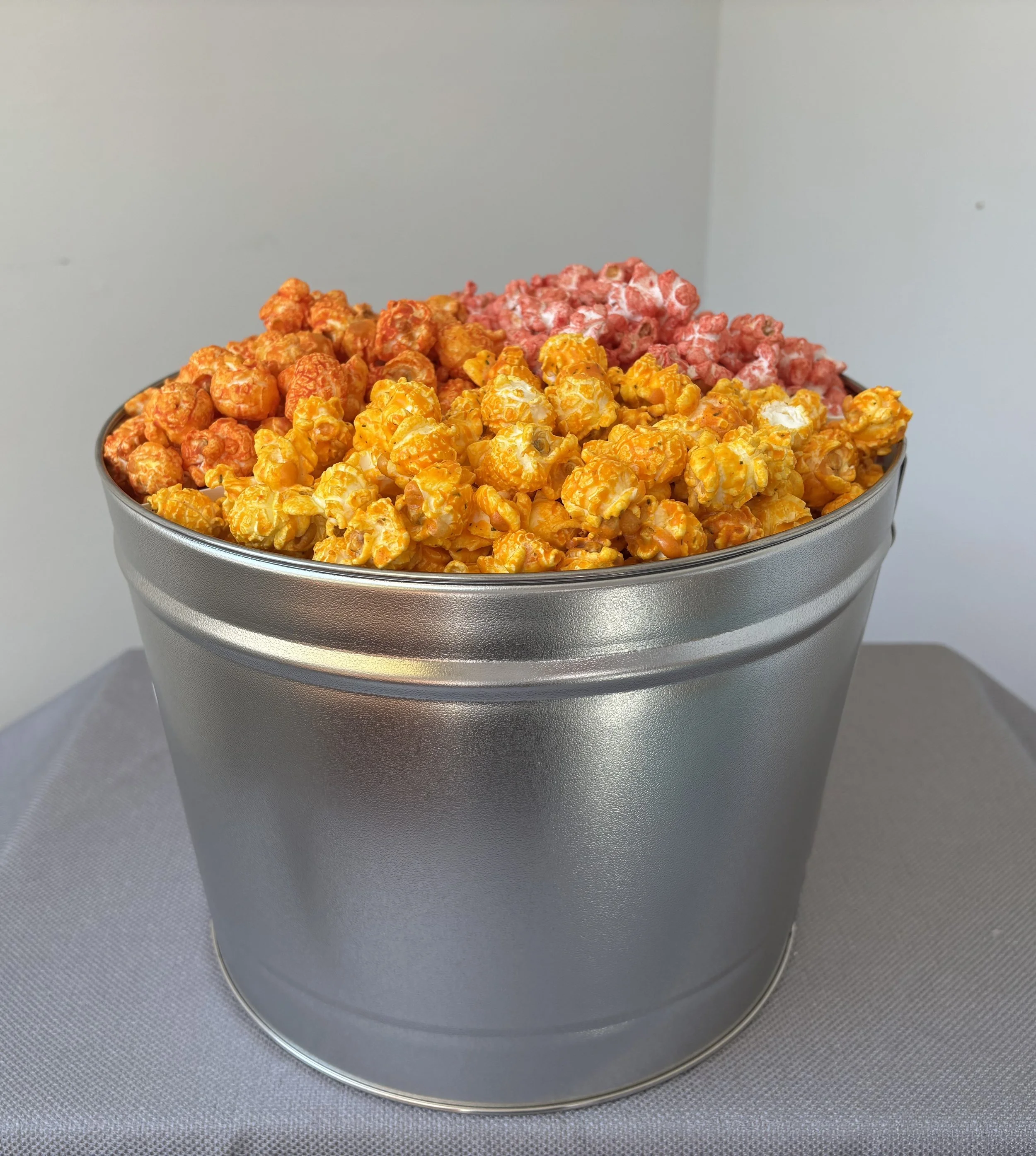 A metal bucket filled with caramel popcorn, with some pink-colored popcorn in the background.