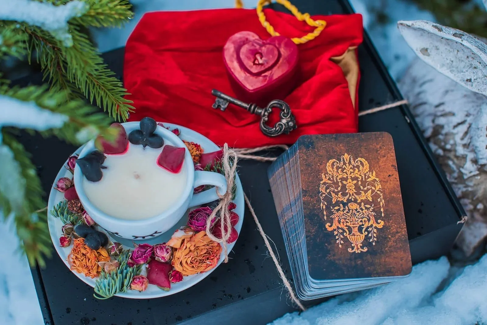 A ceremonial setup with a white candle surrounded by dried flowers on a black surface, a deck of tarot cards with ornate gold design, a red cloth with a vintage key and carved heart piece, and some greenery in the background.