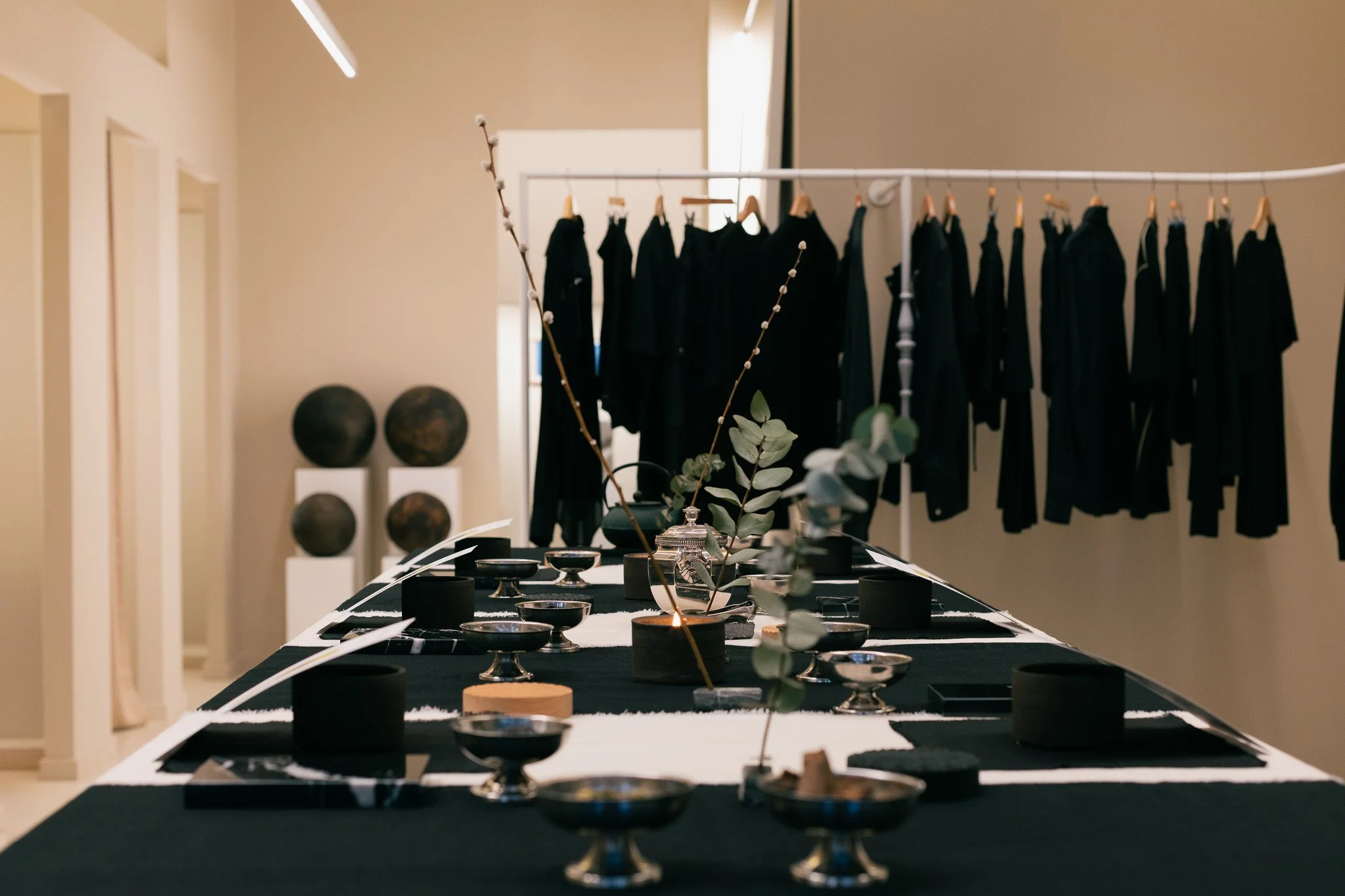 TG Space. Creative encounter. A display table with black and white tablecloths showcasing various black bowls, cups, and decorative items, with a background of black clothing on hangers and two spherical sculptures on stands against a beige wall.