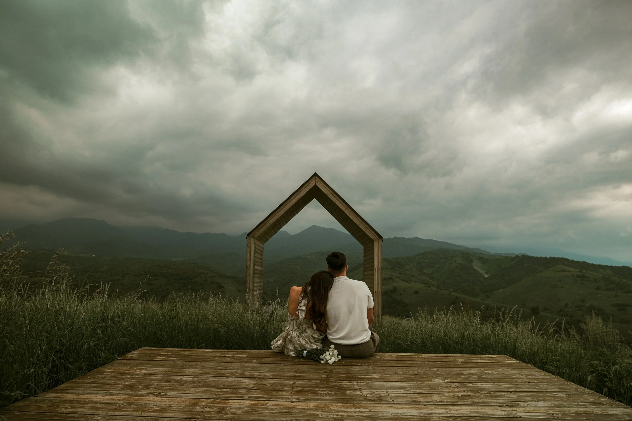 A couple sitting closely on a wooden platform, seen from behind, looking at a scenic mountain landscape under cloudy sky, with an open wooden frame structure behind them.