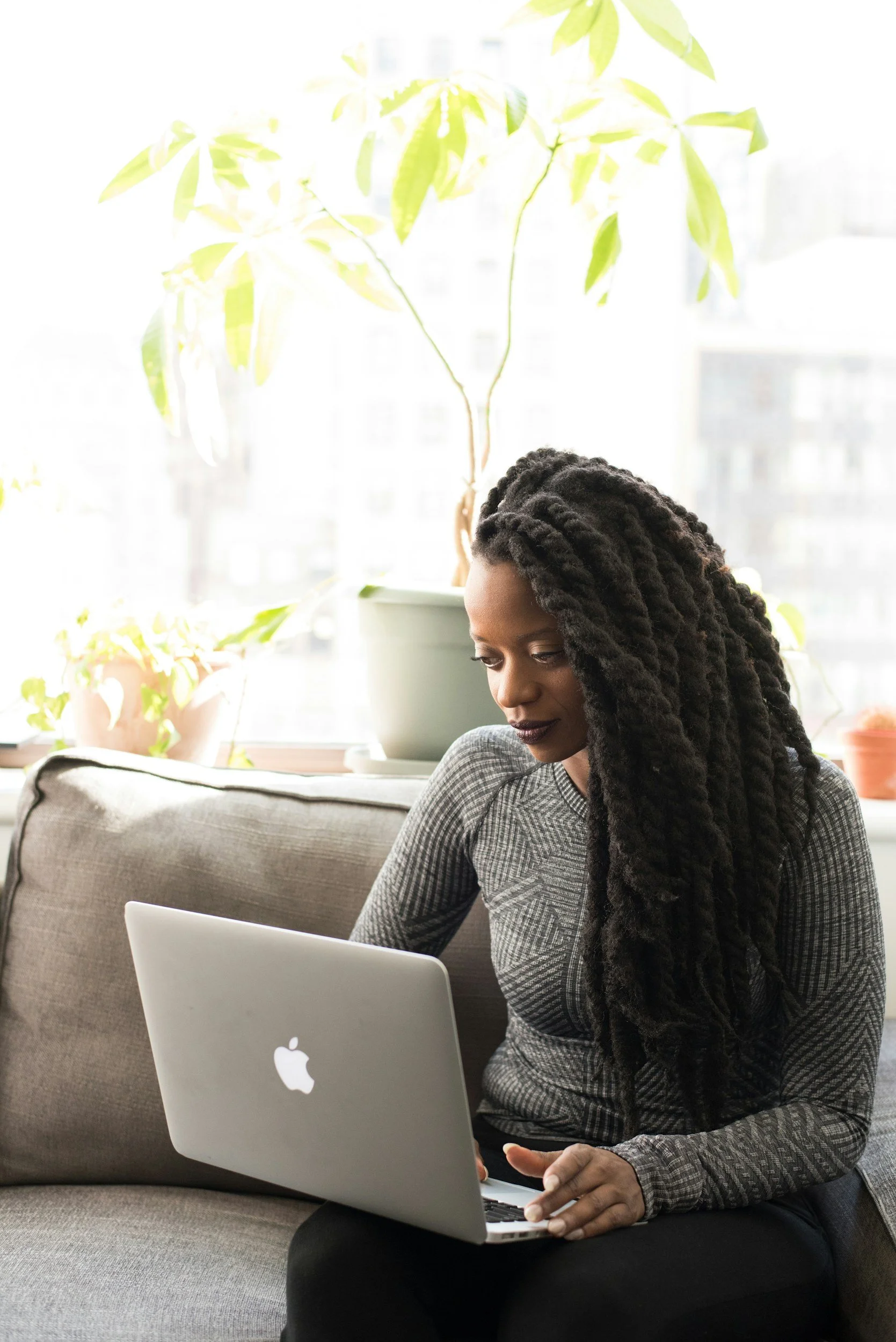 A woman with long dreadlocks working on a silver MacBook laptop while sitting on a gray sofa, with potted plants on the window sill behind her.