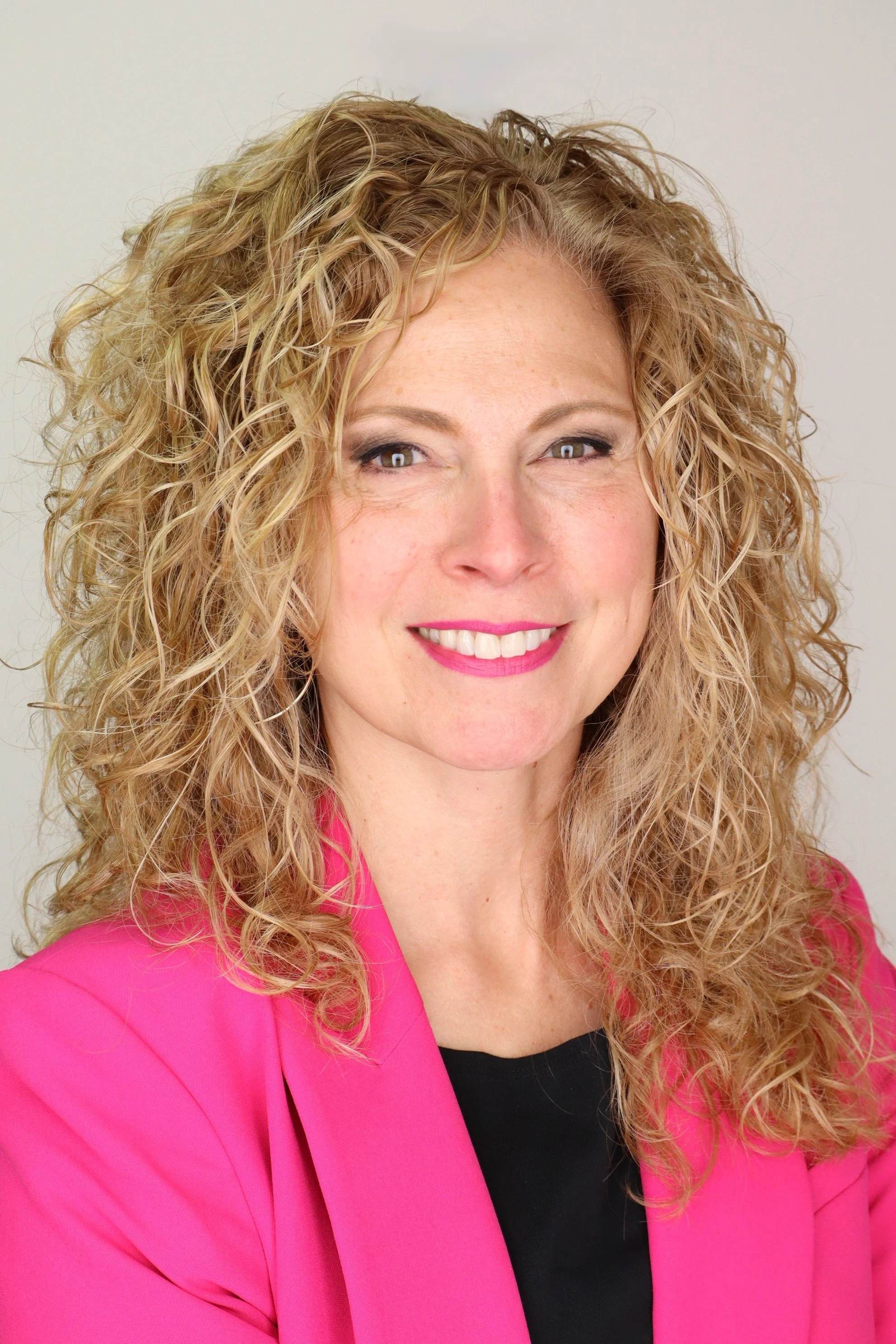 Close-up of a woman with curly blonde hair, wearing a pink blazer and a black top, smiling slightly against a plain light background.