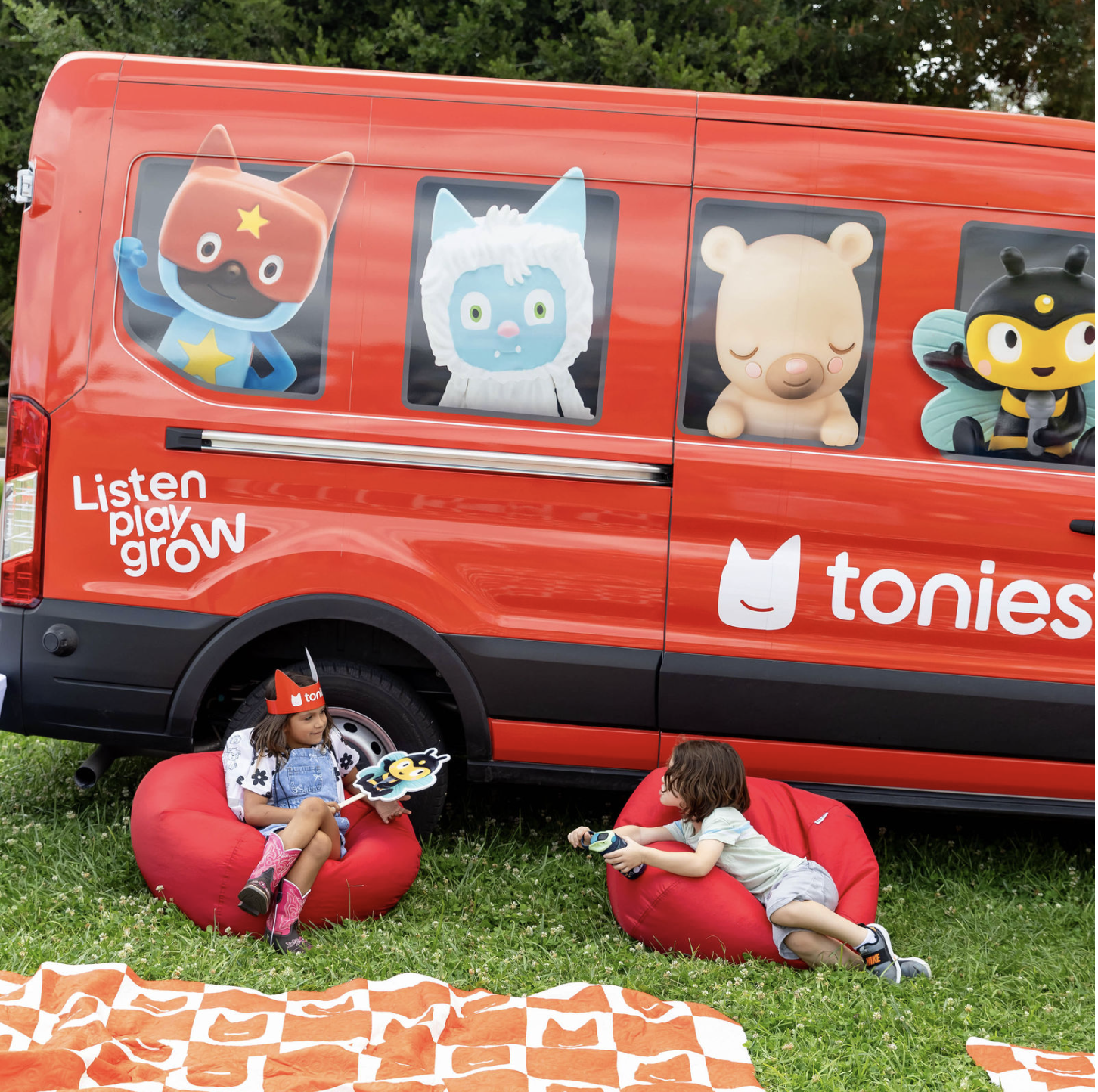 Red speaker with a black stylized cat face made of holes, two black tail-like antennae on top, positioned on a wooden surface. Next to it are two toy figures: one in a red costume with cat ears and a smiling face, and another resembling a brown owl with large eyes holding a stick. In the background, a white box with the red logo and text 'tonies' and a smiling cat face.