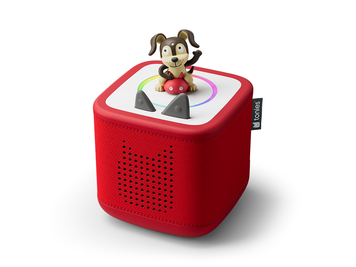 Red speaker with a black stylized cat face made of holes, two black tail-like antennae on top, positioned on a wooden surface. Next to it are two toy figures: one in a red costume with cat ears and a smiling face, and another resembling a brown owl with large eyes holding a stick. In the background, a white box with the red logo and text 'tonies' and a smiling cat face.