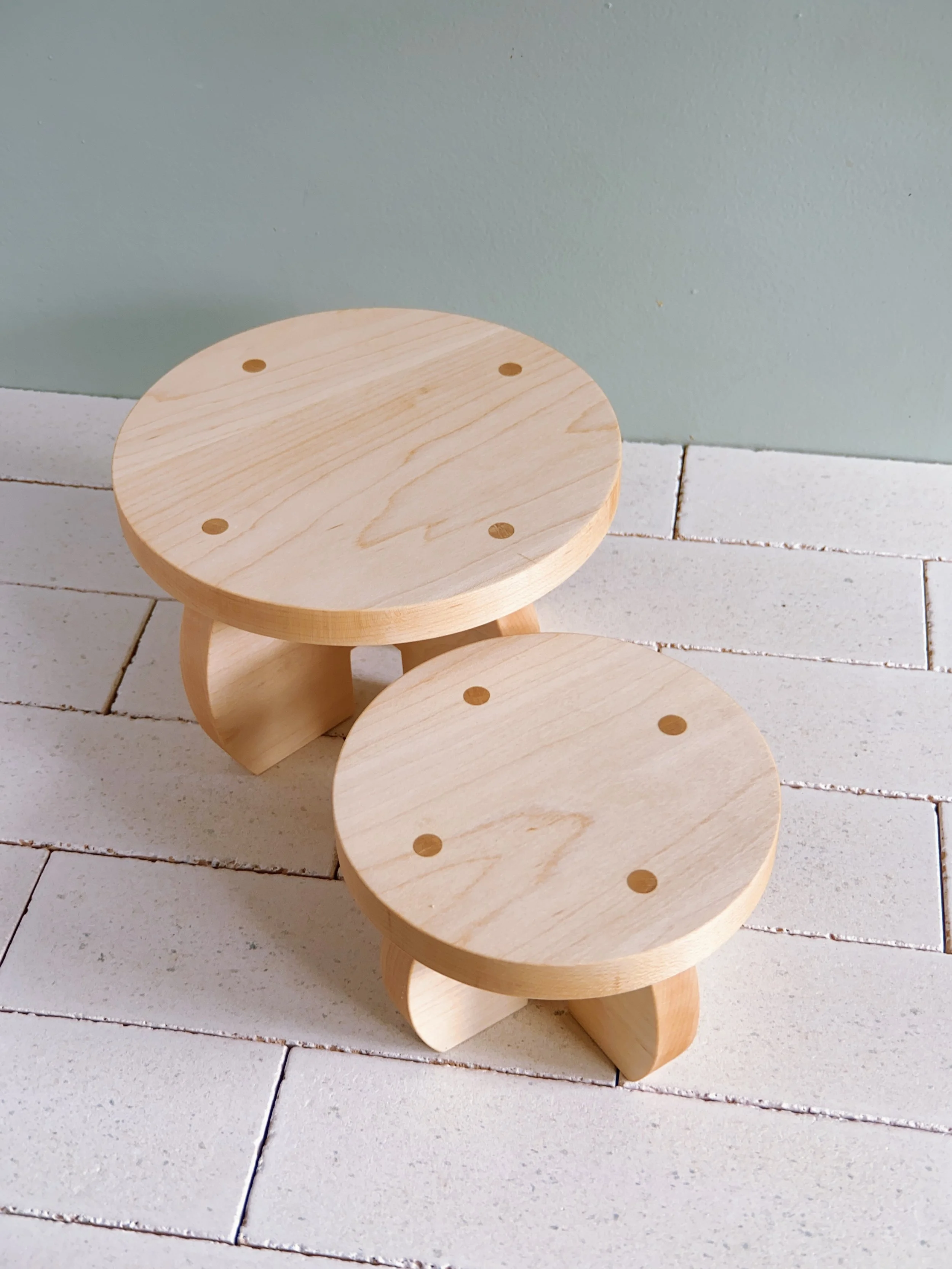 Two small round wooden plant stands with flat tops and curved legs, placed on a white tiled countertop against a plain green wall.