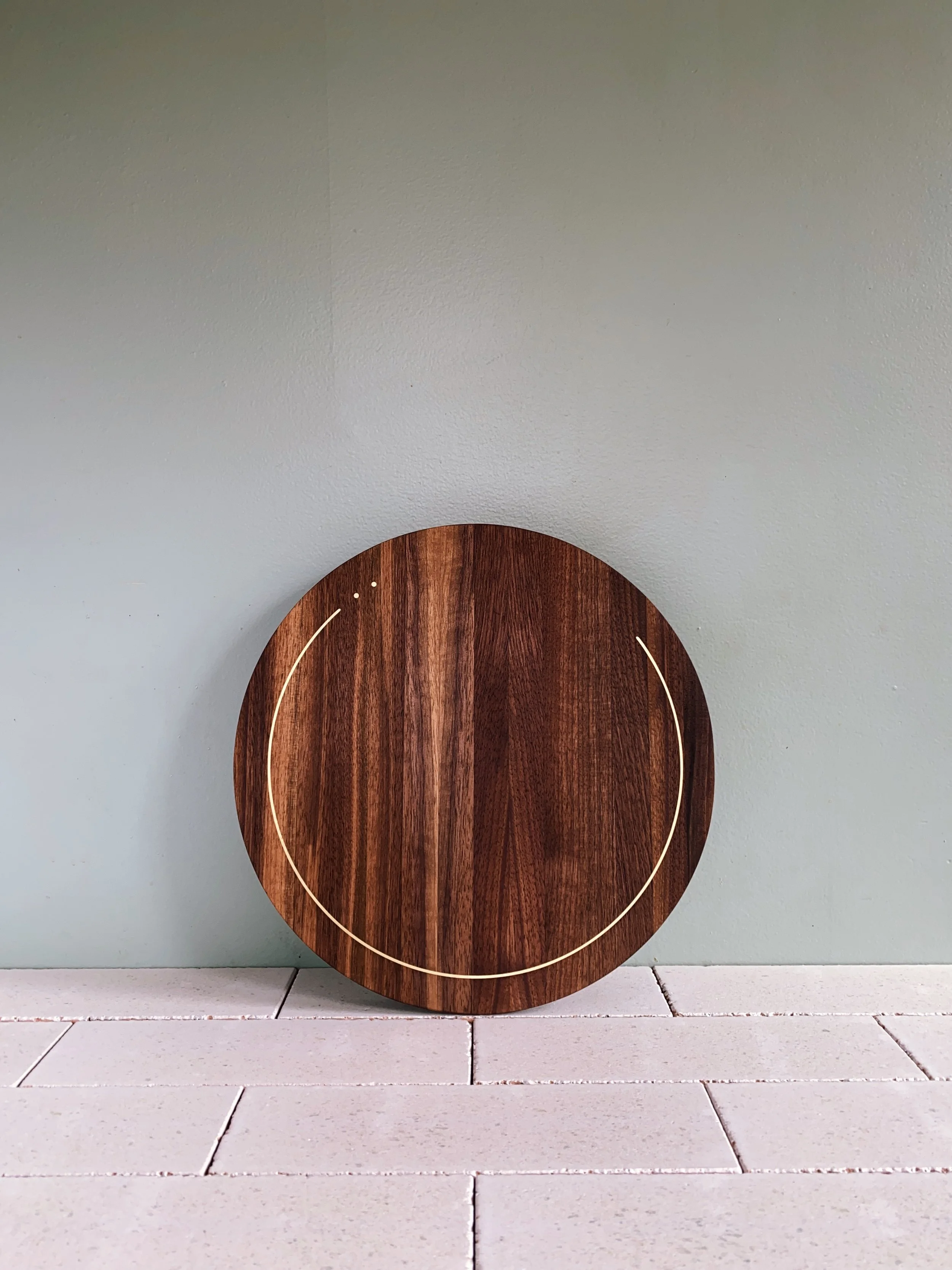 Round wooden wall clock with minimalistic design, no numbers, mounted on a pale green wall above white tiled countertop.