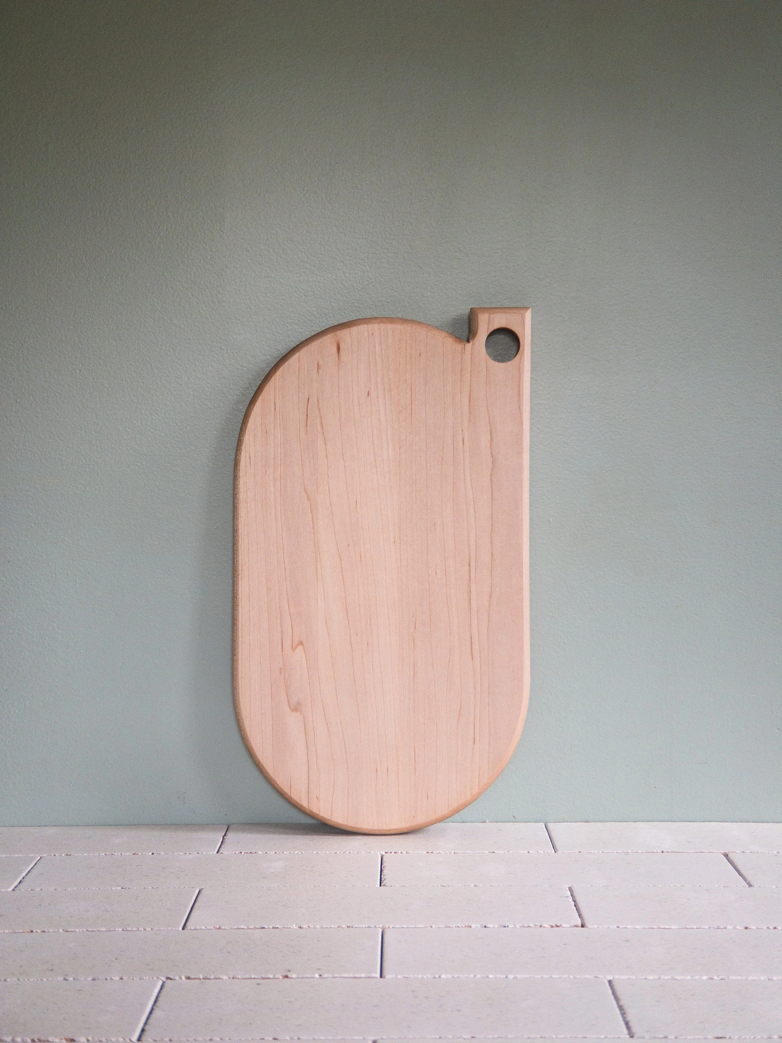 Wooden cutting board with a hole in the top right corner, placed against a green wall and on a tiled countertop.
