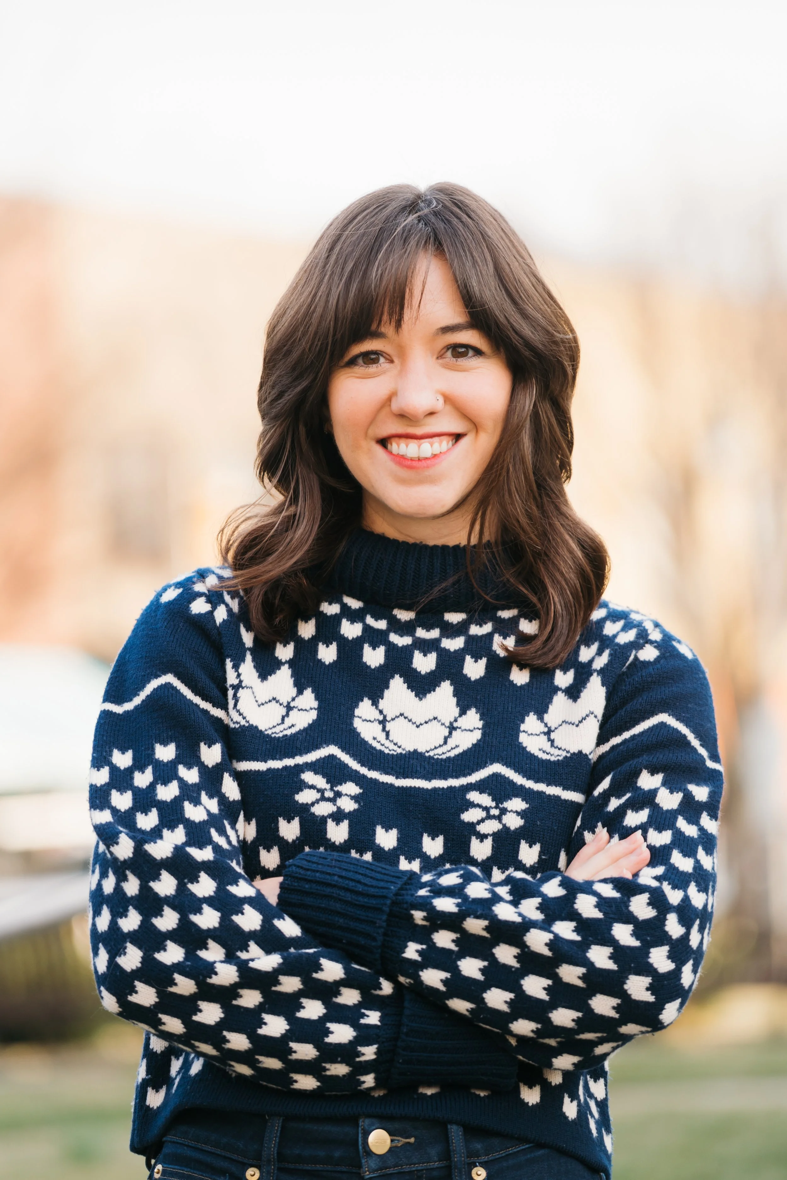 A woman with shoulder-length brown hair smiling outdoors, wearing a navy blue and white patterned sweater.