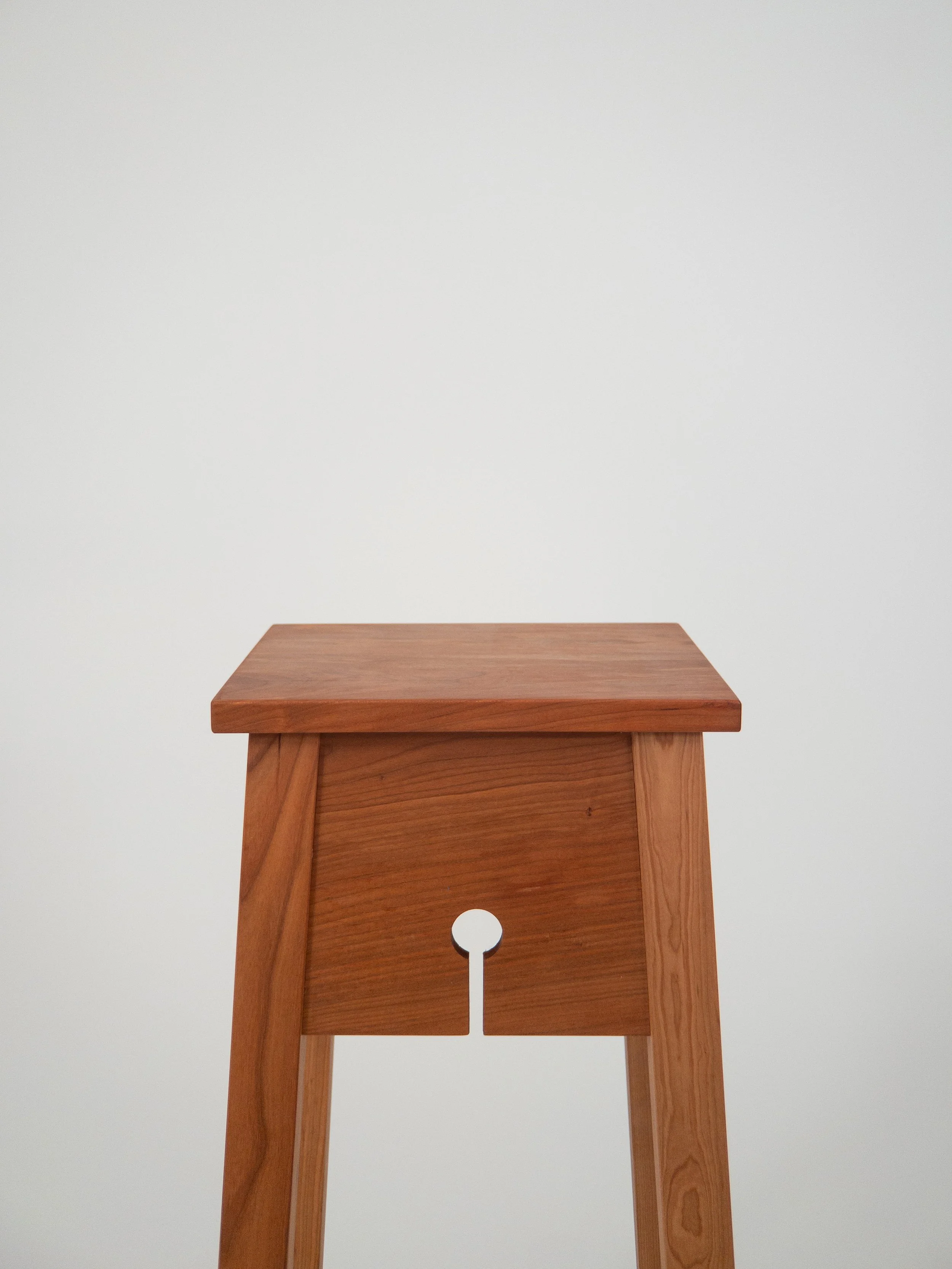 A wooden pedestal with four legs and a keyhole design in the center of its side panel, set against a plain light-colored wall.