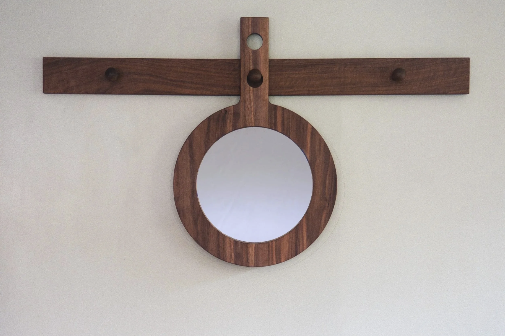 A round mirror with a dark walnut wooden frame hanging from a dark walnut wooden pegboard mounted on a light blue wall.